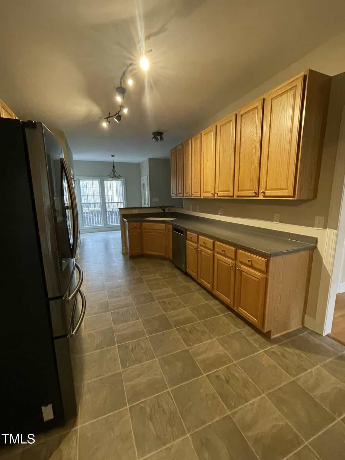 Long kitchen with tiled floor, black refrigerator, dishwasher, and wooden cabinets. Track lighting above. Large window in the background lets in natural light. Neutral colors throughout.