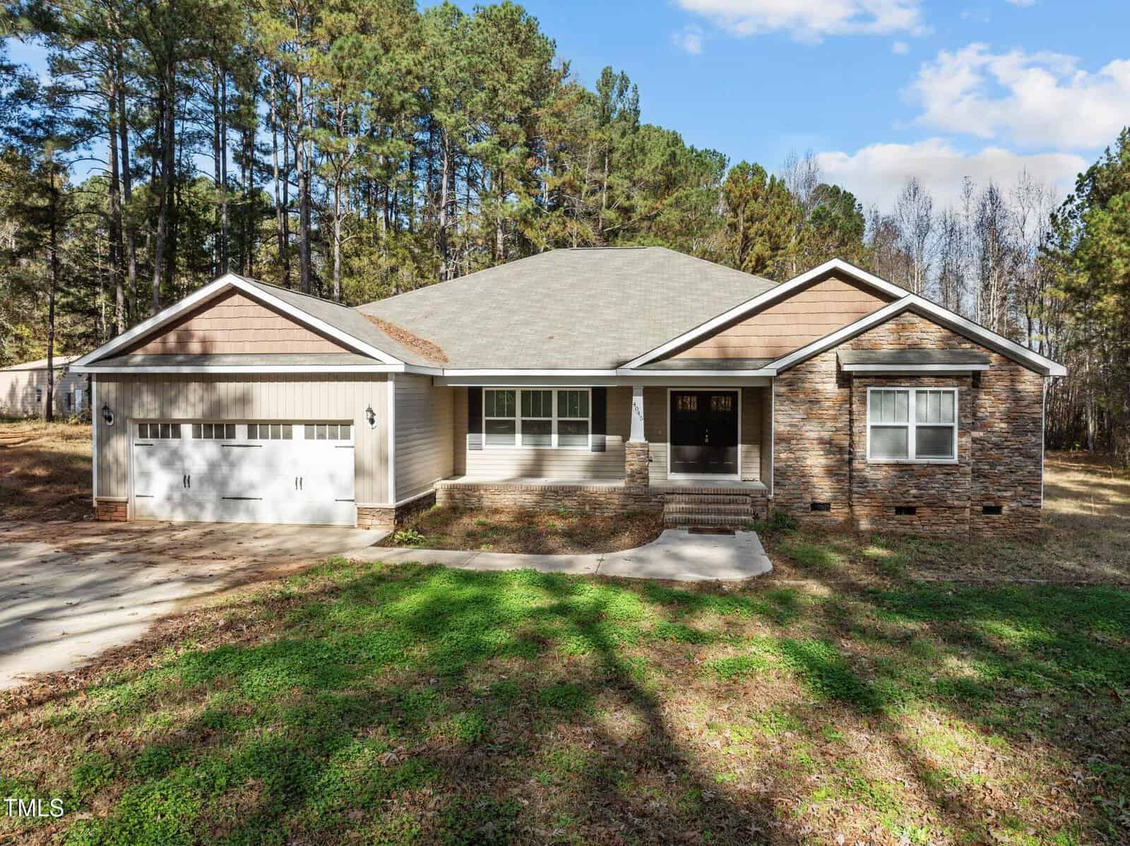 A single-story house with a light brown exterior, featuring a two-car garage on the left and a stone facade on the right. The home is surrounded by trees, and a concrete driveway leads to the entrance. The sky is clear with a few clouds.