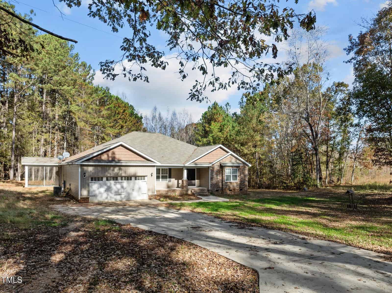 Single-story house with a gray roof and beige siding, surrounded by trees. A paved driveway leads to a two-car garage. The yard is dotted with autumn leaves. The sky is partly cloudy, with patches of blue visible.