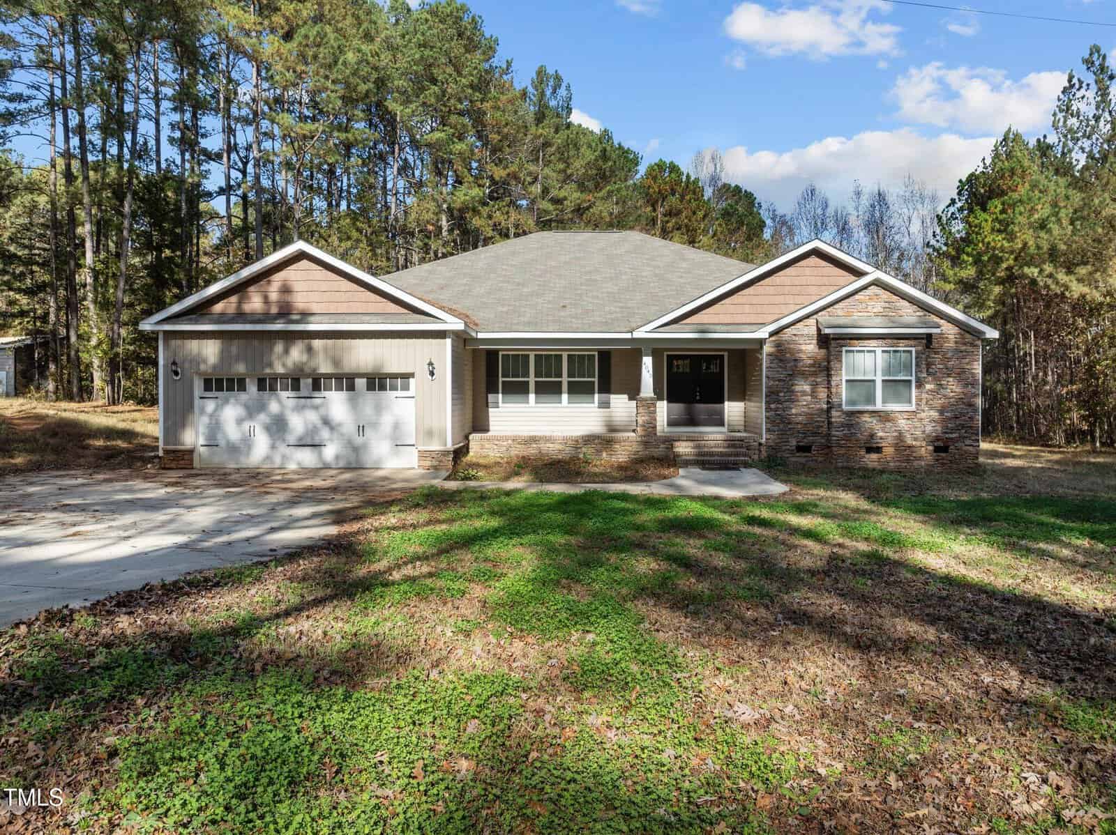 A single-story house with a brick and siding exterior is surrounded by a wooded area. The house features a two-car garage and a front porch. The sky is clear and sunny, casting shadows on the driveway and lawn.