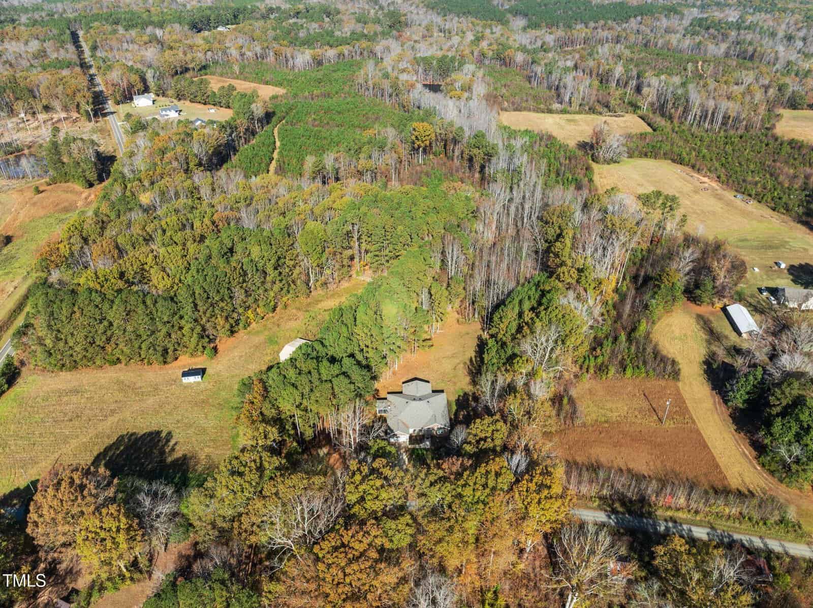 Aerial view of a rural landscape with a house surrounded by trees in the center. The area features a mix of wooded regions and open fields. A road runs near the bottom edge, and a few smaller structures are scattered throughout the scene.