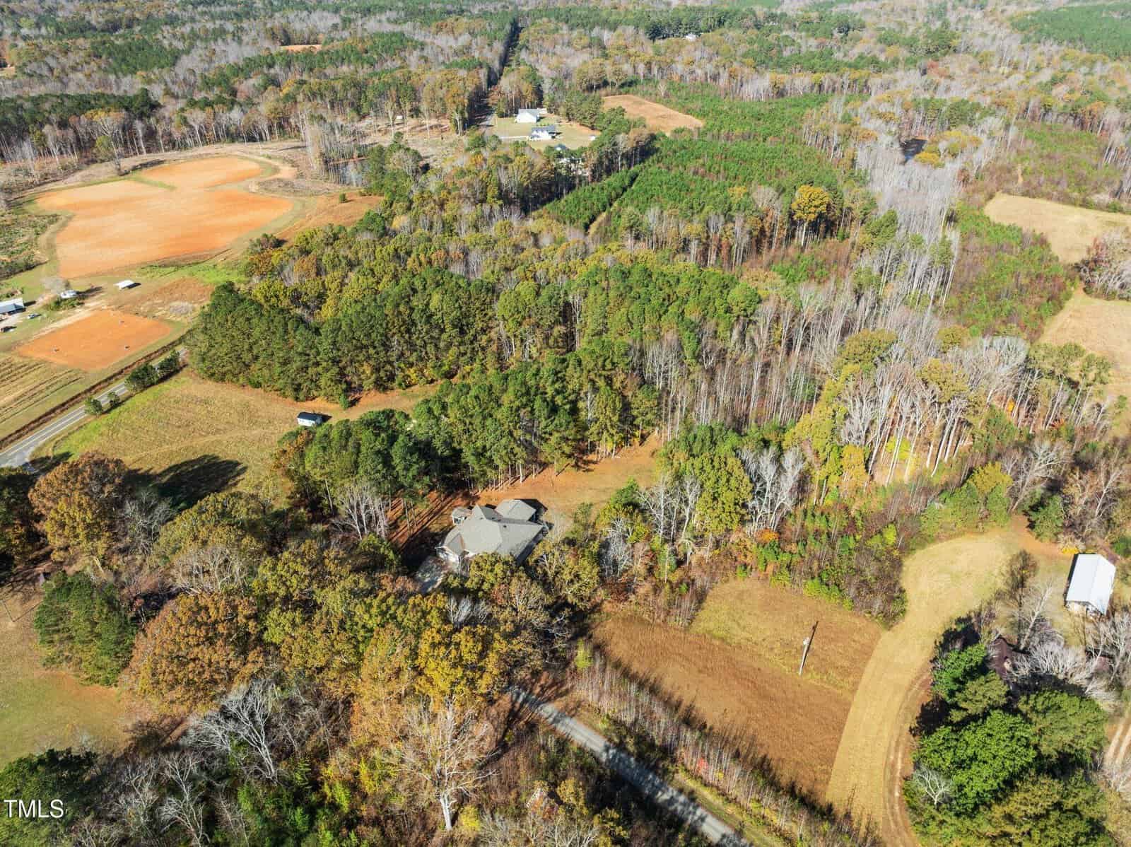 Aerial view of a rural landscape with a mix of forests and fields. A house is partially visible among trees in the center, while open fields and patches of forest surround it. Several dirt roads and small structures are scattered throughout the area.