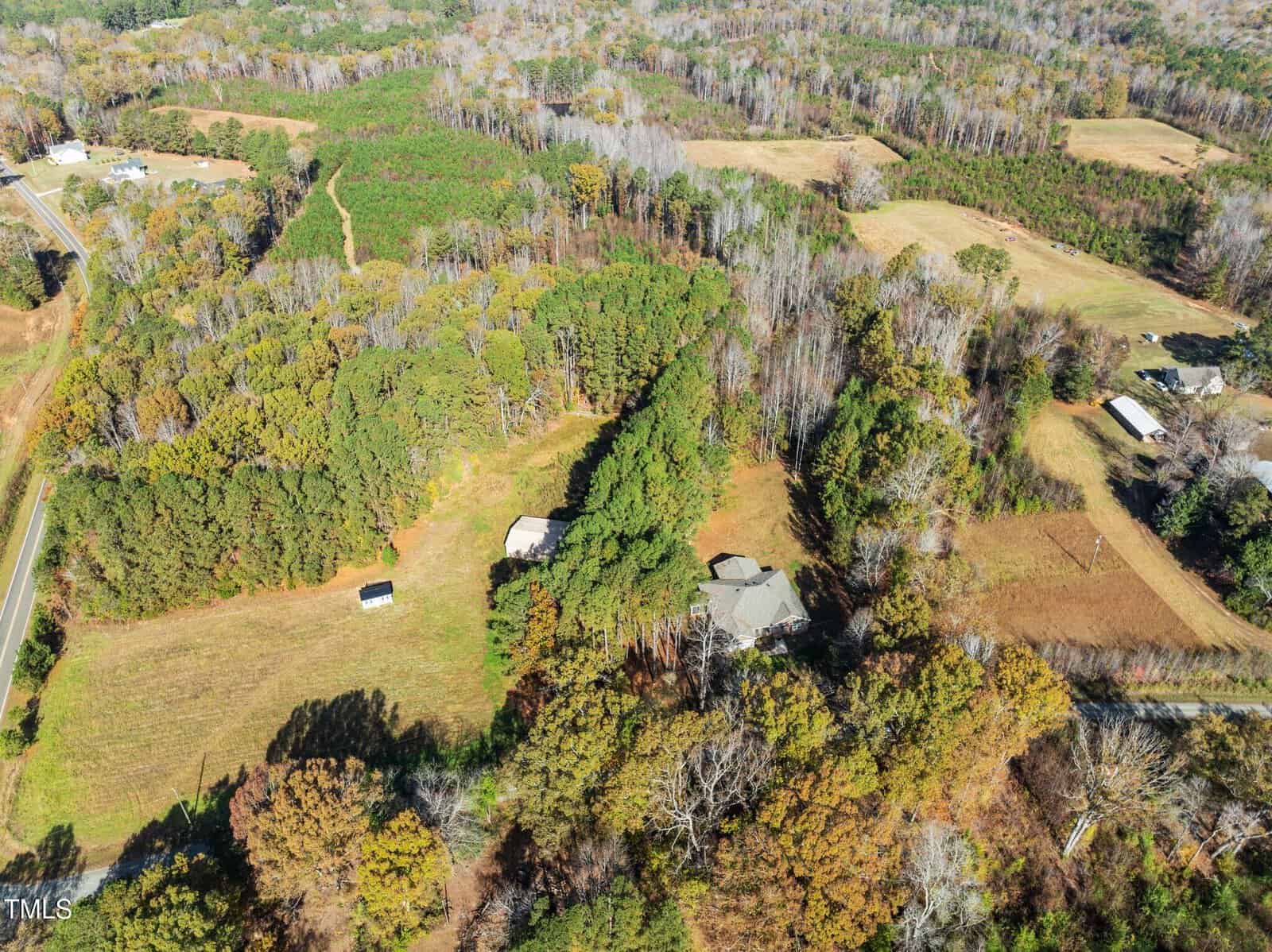 Aerial view of a rural landscape featuring dense wooded areas and open fields. Several houses are scattered throughout, with a road running alongside. The scene is surrounded by trees with autumn foliage.