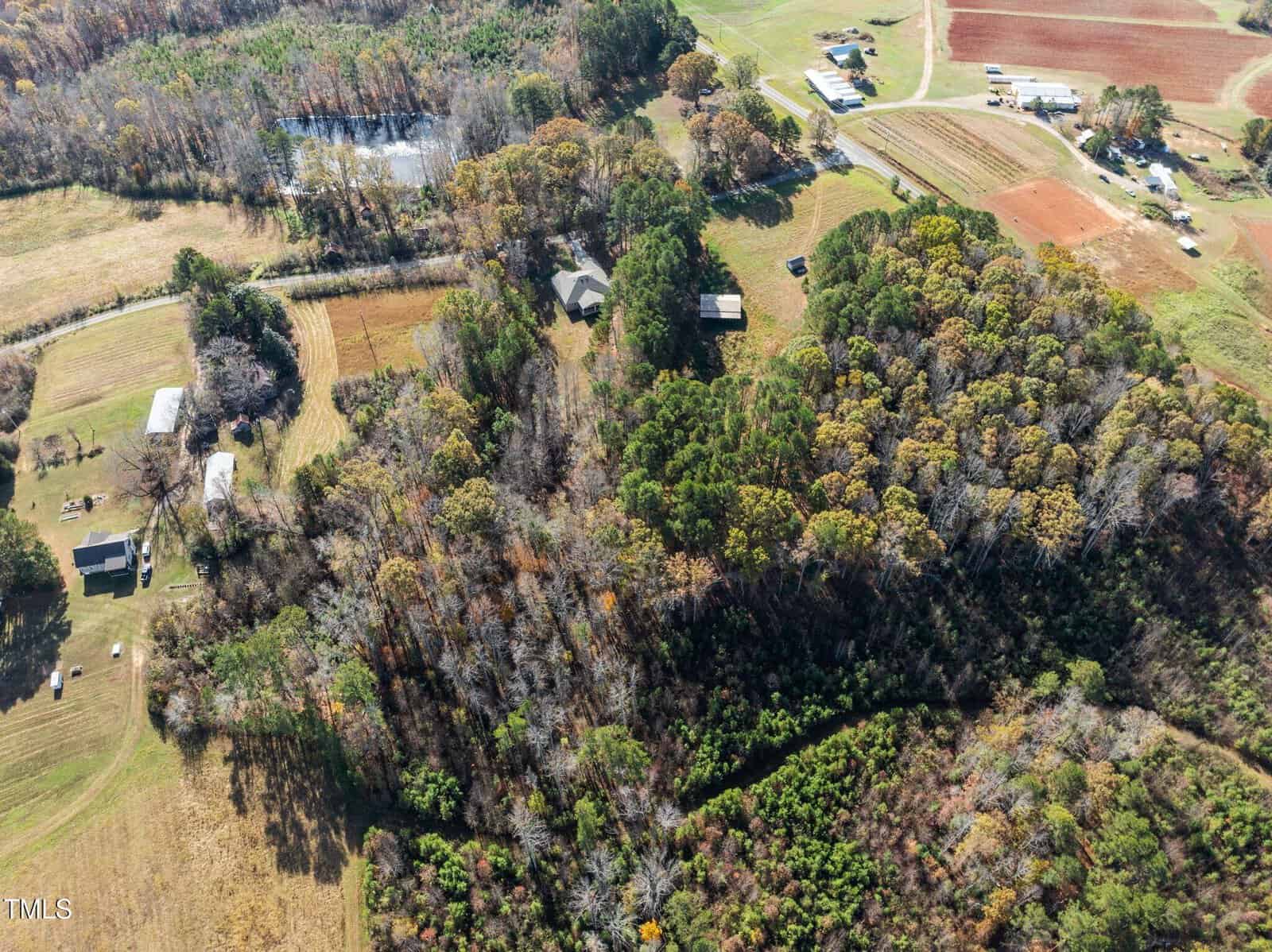 Aerial view of a rural landscape featuring dense patches of trees, open fields, and scattered buildings. There are several roads and a pond surrounded by foliage in the upper left corner.