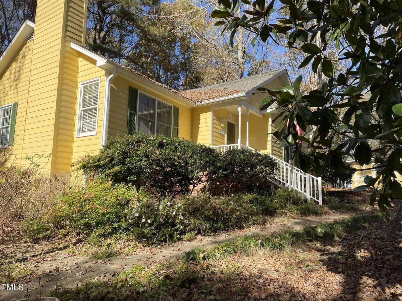 A yellow house with white trim and a small porch, surrounded by bushes and trees. The walkway is lined with fallen leaves, and sunlight filters through the branches above.