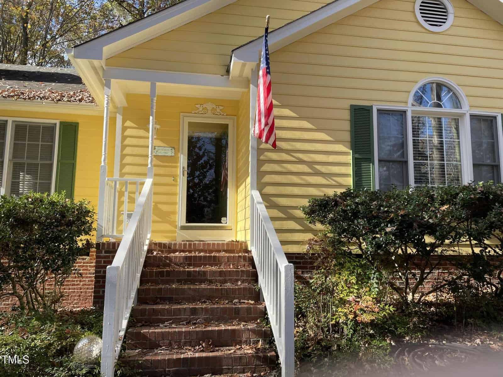 A yellow house with white trim, a green shutter, and an American flag mounted near the front porch. Brick steps with white railings lead to the front door. Shrubs and trees are in the yard.