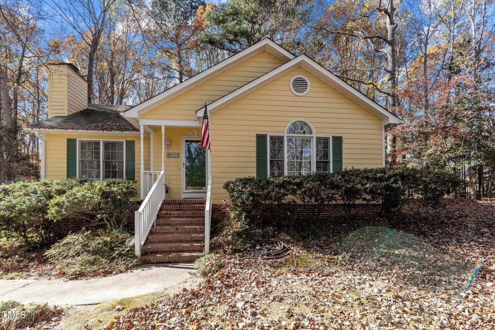 A small yellow house with green shutters and a brick foundation, featuring a front porch with steps, an American flag, and surrounded by bushes and autumn trees. Fallen leaves cover the yard and walkway.