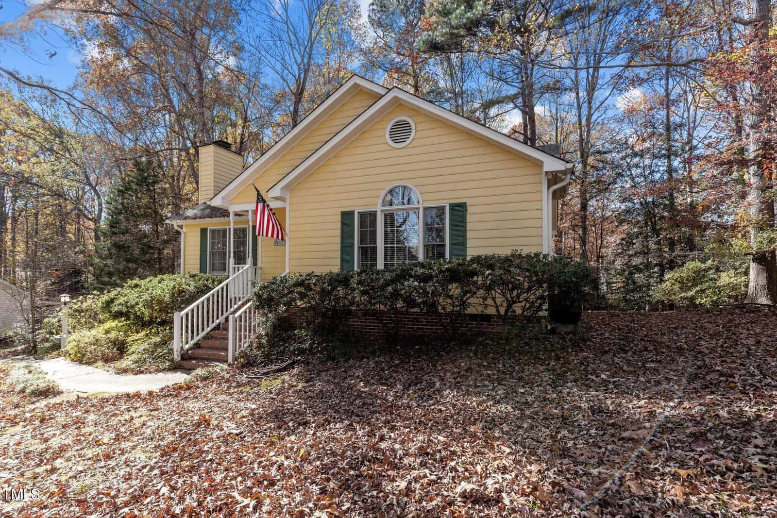 A yellow single-story house with green shutters and a front porch, displaying an American flag. The yard is covered in fallen leaves and surrounded by tall trees with autumn foliage.