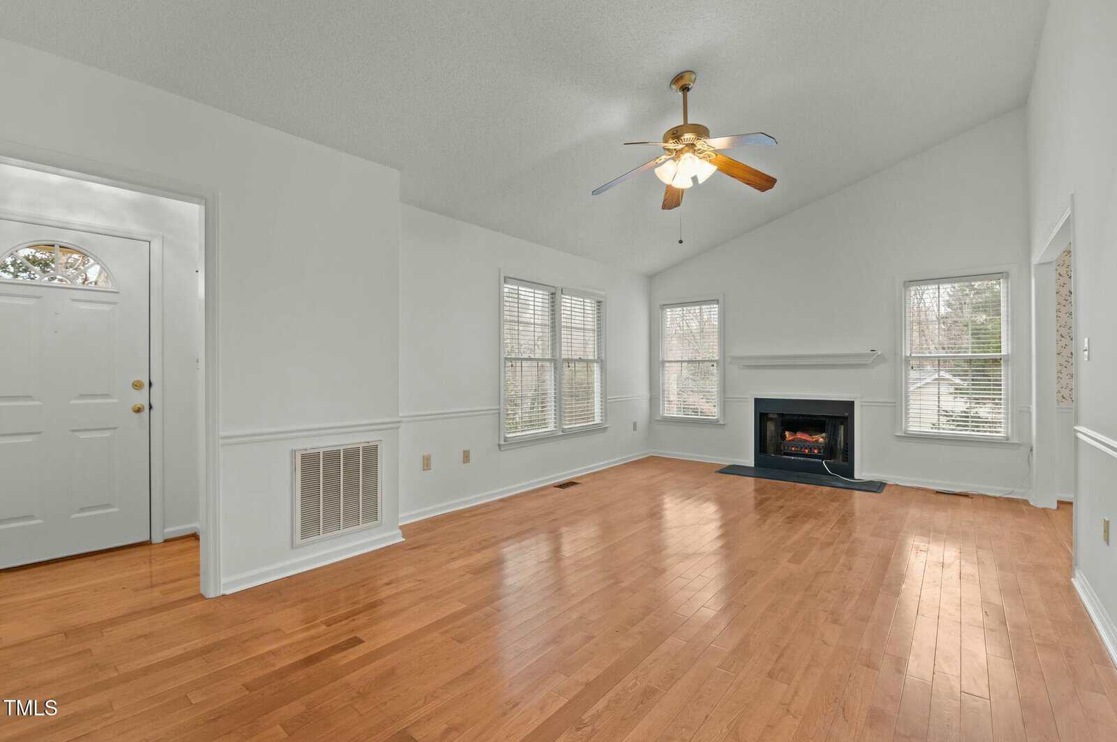A spacious living room with light wood floors, white walls, a ceiling fan with lights, a fireplace, several windows, and a white front door; the room is empty and well-lit with natural light.