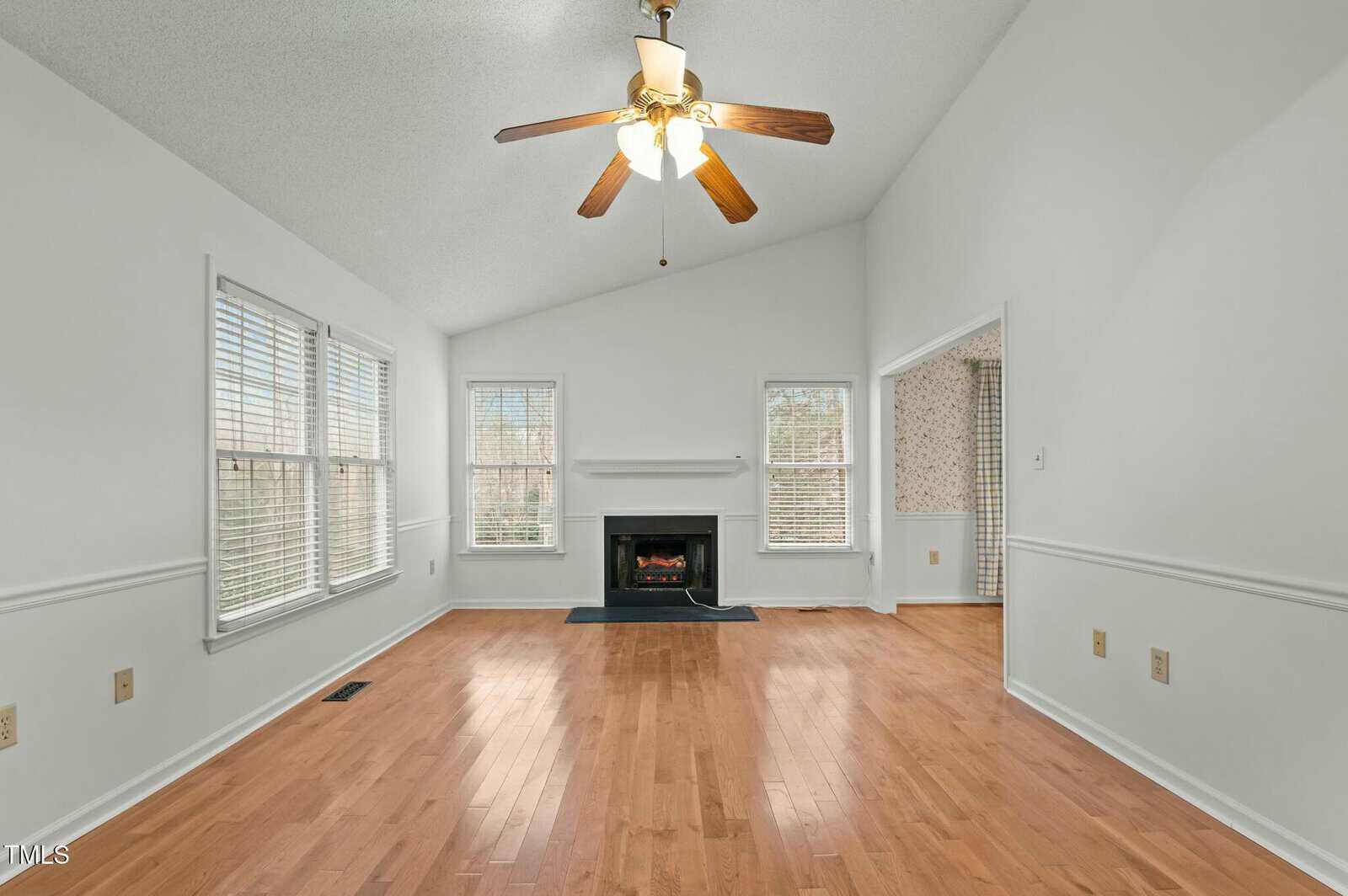 Bright living room with wood floors, white walls, a ceiling fan, large windows, a black fireplace, and an open doorway leading to another room.