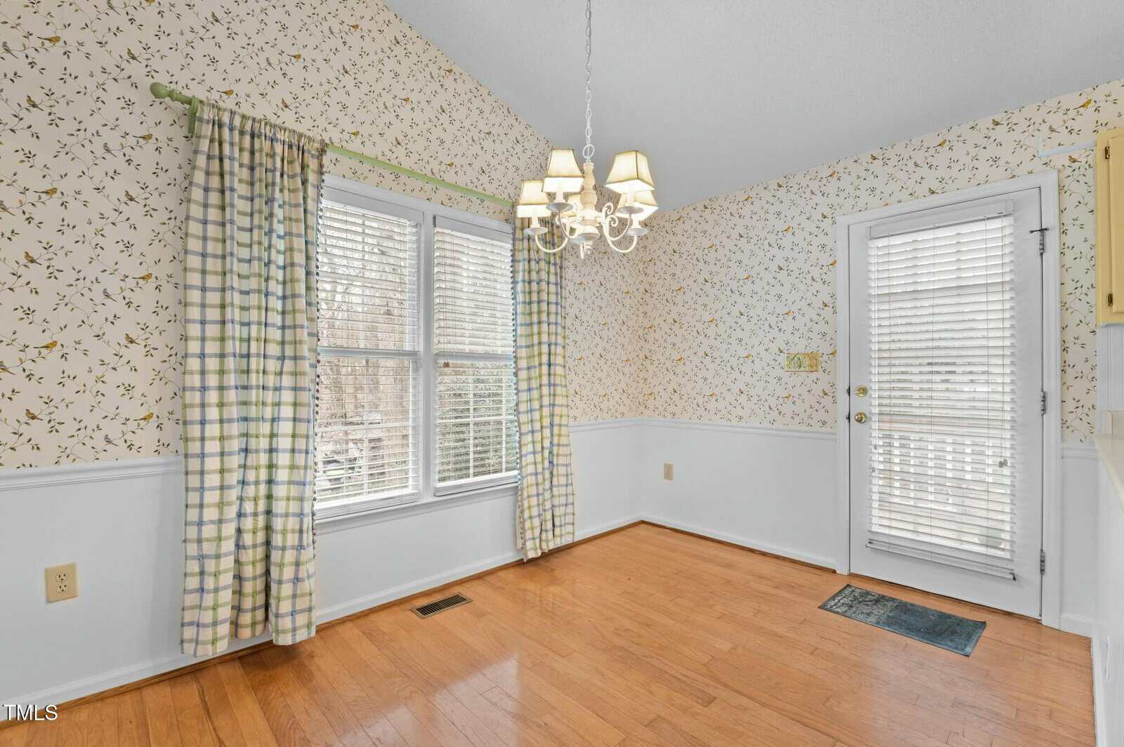 A bright dining area with plaid curtains, floral wallpaper, a white chandelier, wood floor, large windows, and a glass door with blinds leading outside. A small mat is by the door.