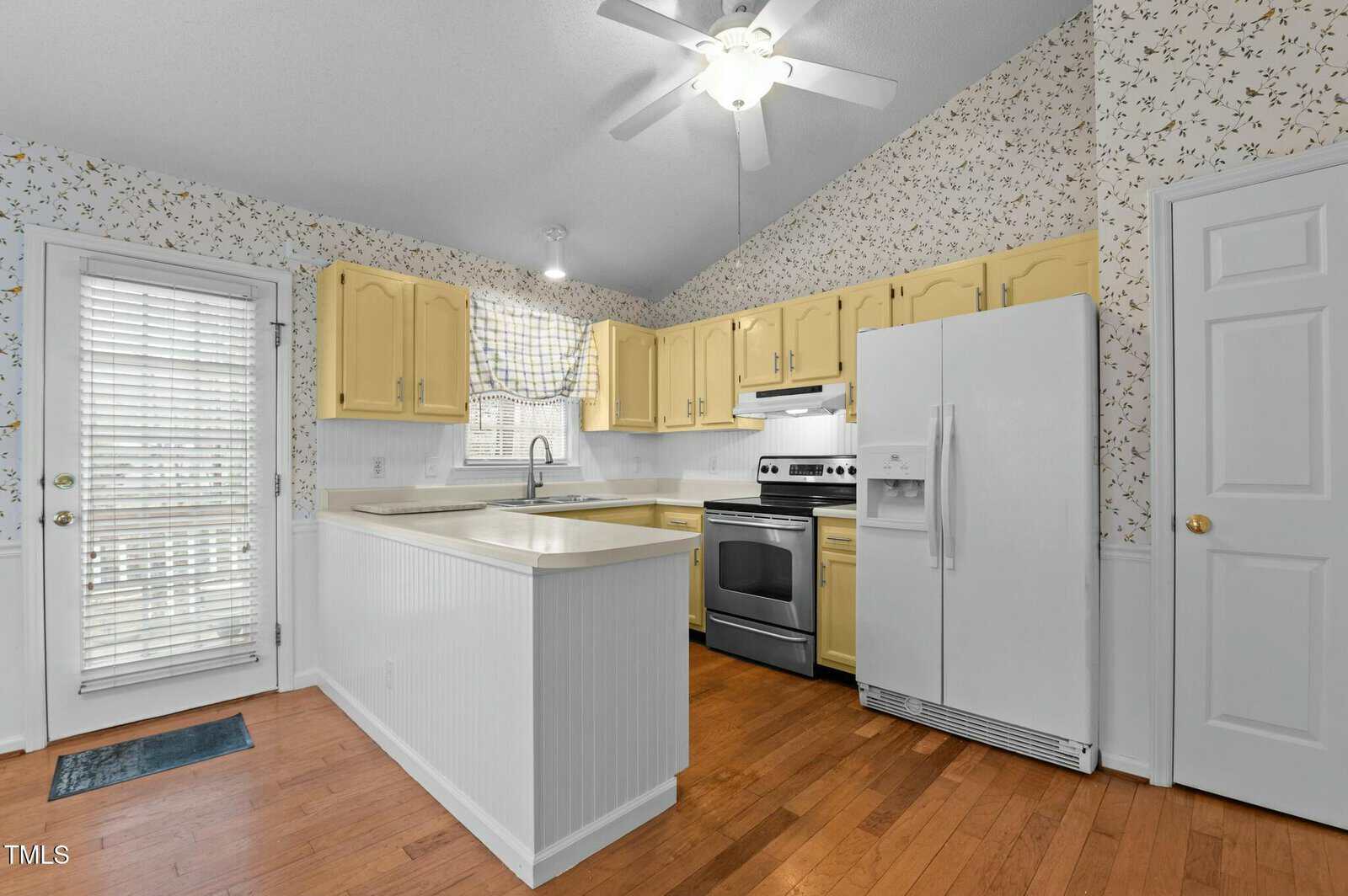 A kitchen with yellow cabinets, white countertops, stainless steel stove, white refrigerator, floral wallpaper, wood floors, and a window with a checkered valance above the sink. A door with blinds is on the left.