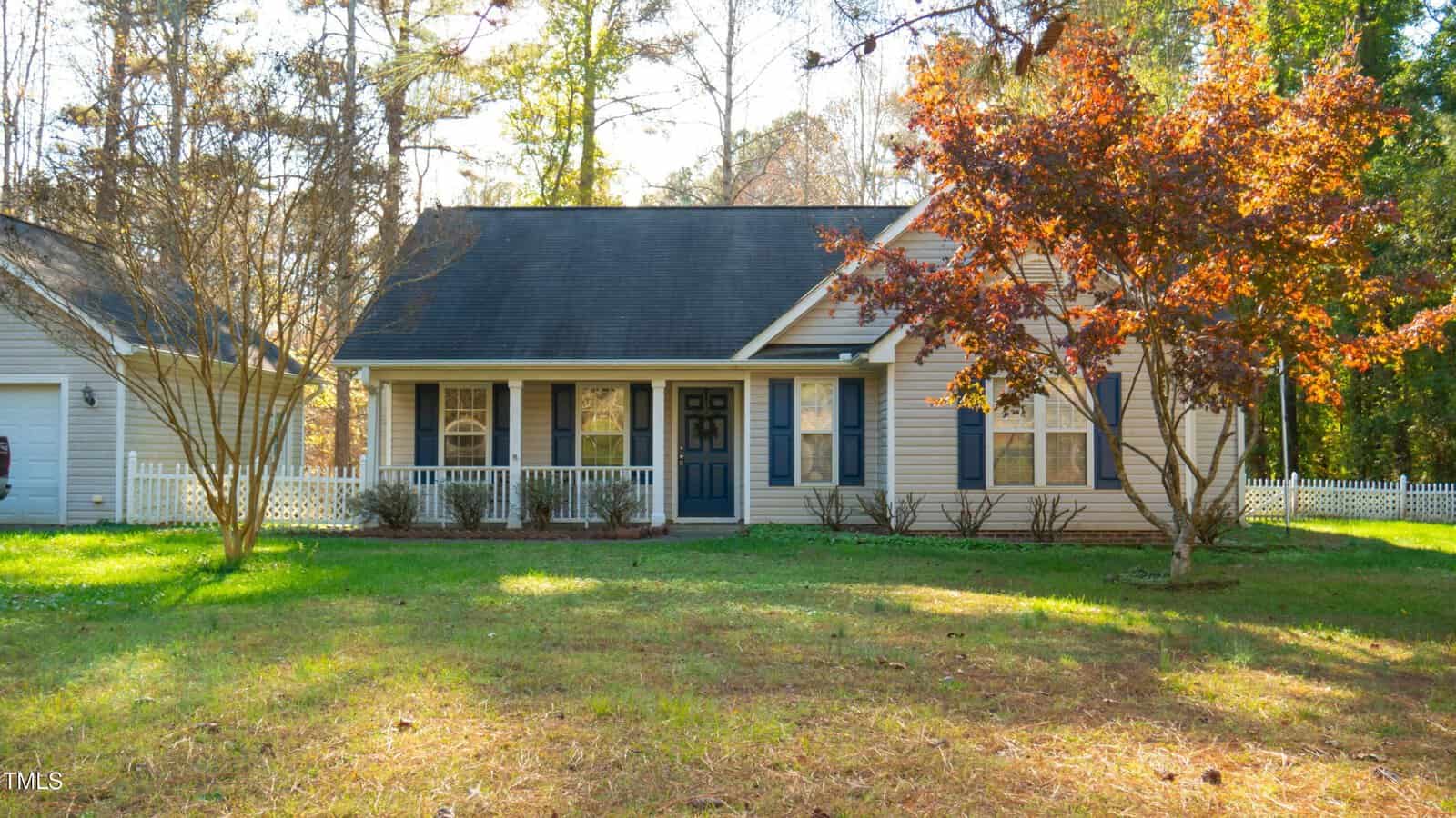 A quaint suburban house with a dark roof and light siding is surrounded by trees. The front yard has a few bare trees, and a vibrant red-leaved tree. A white picket fence runs along the property, and the grass is lush and green.