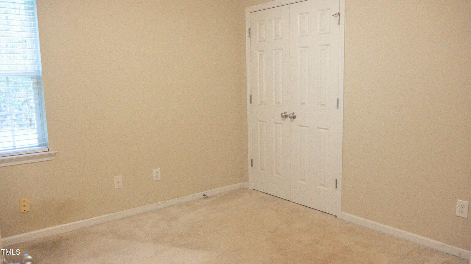 A small, empty room with beige walls and carpet. There is a closed white double door closet on the right and a window with blinds on the left. The room is well-lit and has electrical outlets visible on the walls.