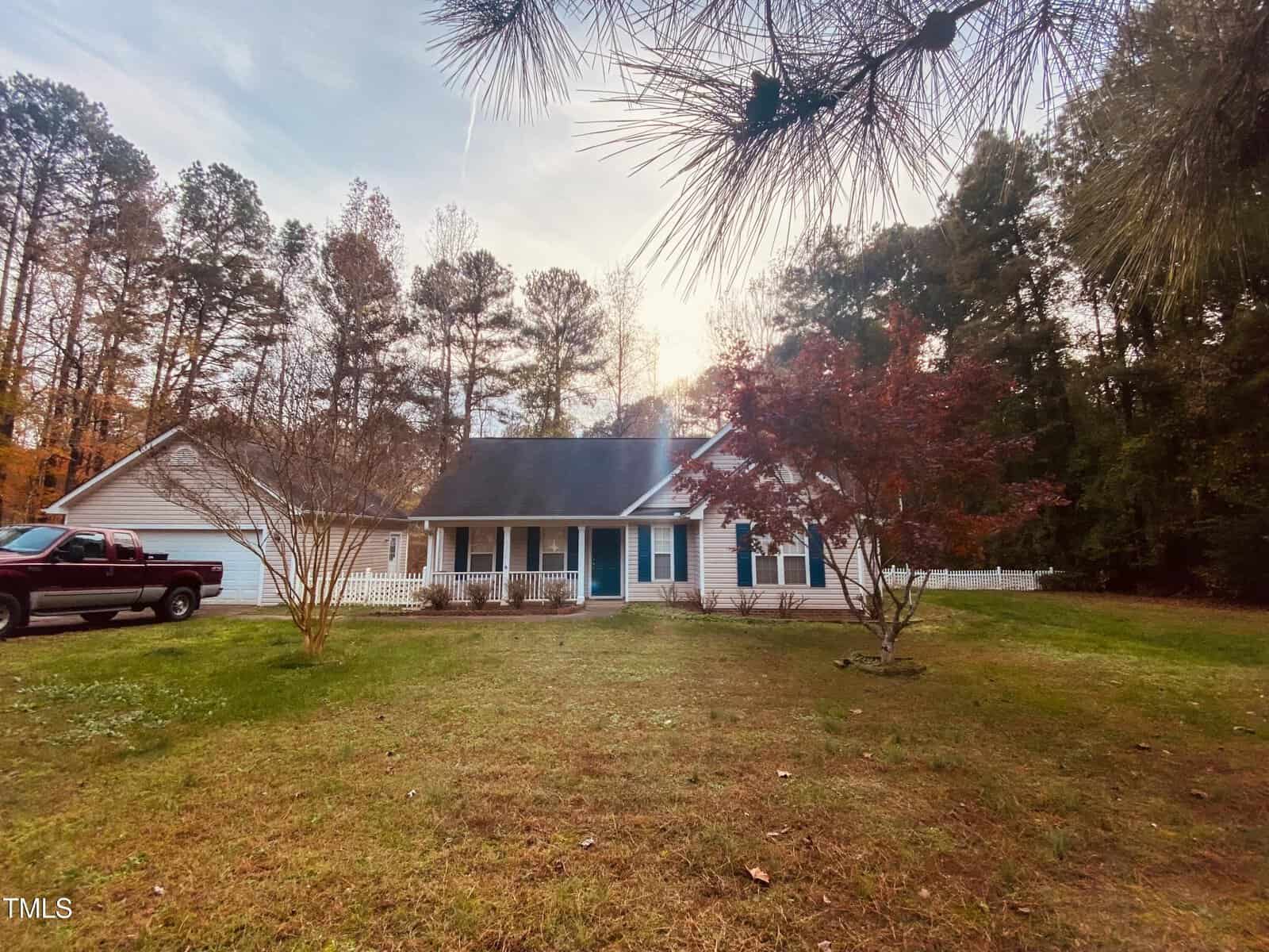 A single-story house with a porch sits amidst a grassy yard. A red truck is parked on the left side. Tall trees with autumn leaves surround the property. The sky is partly cloudy.