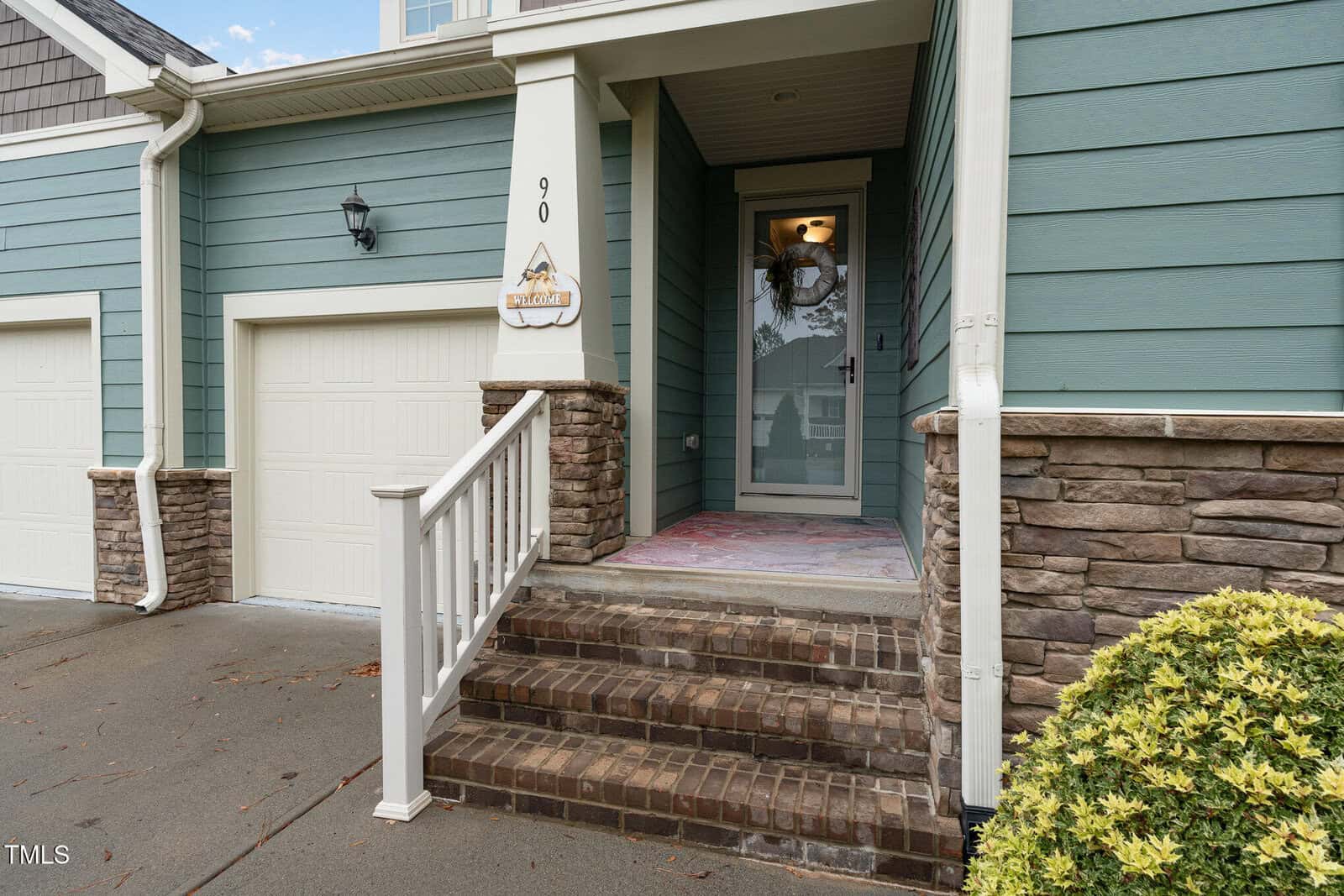 Front porch of a house with blue siding and stone accents, white garage door, brick steps, and a glass front door. A welcome sign hangs by the house number 90. Shrubs and a wet driveway are visible.