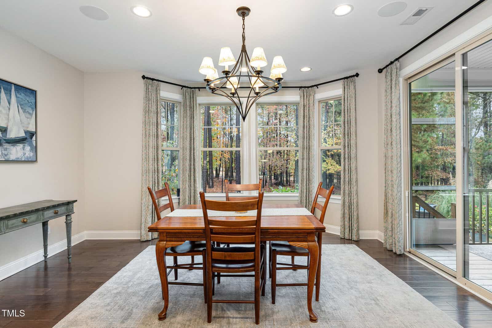 A dining room with a wooden table and six chairs sits on a light rug. A chandelier hangs above. Large windows with curtains reveal a wooded view. A painting and a small table are on the left wall. Hardwood floors are visible.