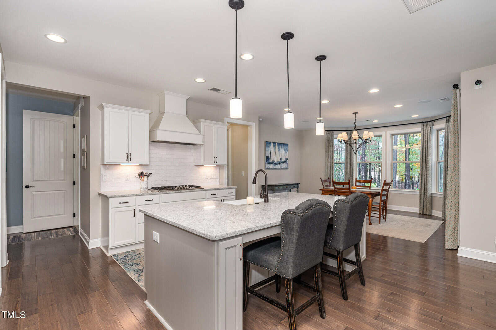 Modern kitchen with a large island, gray upholstered chairs, white cabinets, and stainless steel appliances. Dining area with chandelier and large windows is visible in the background.