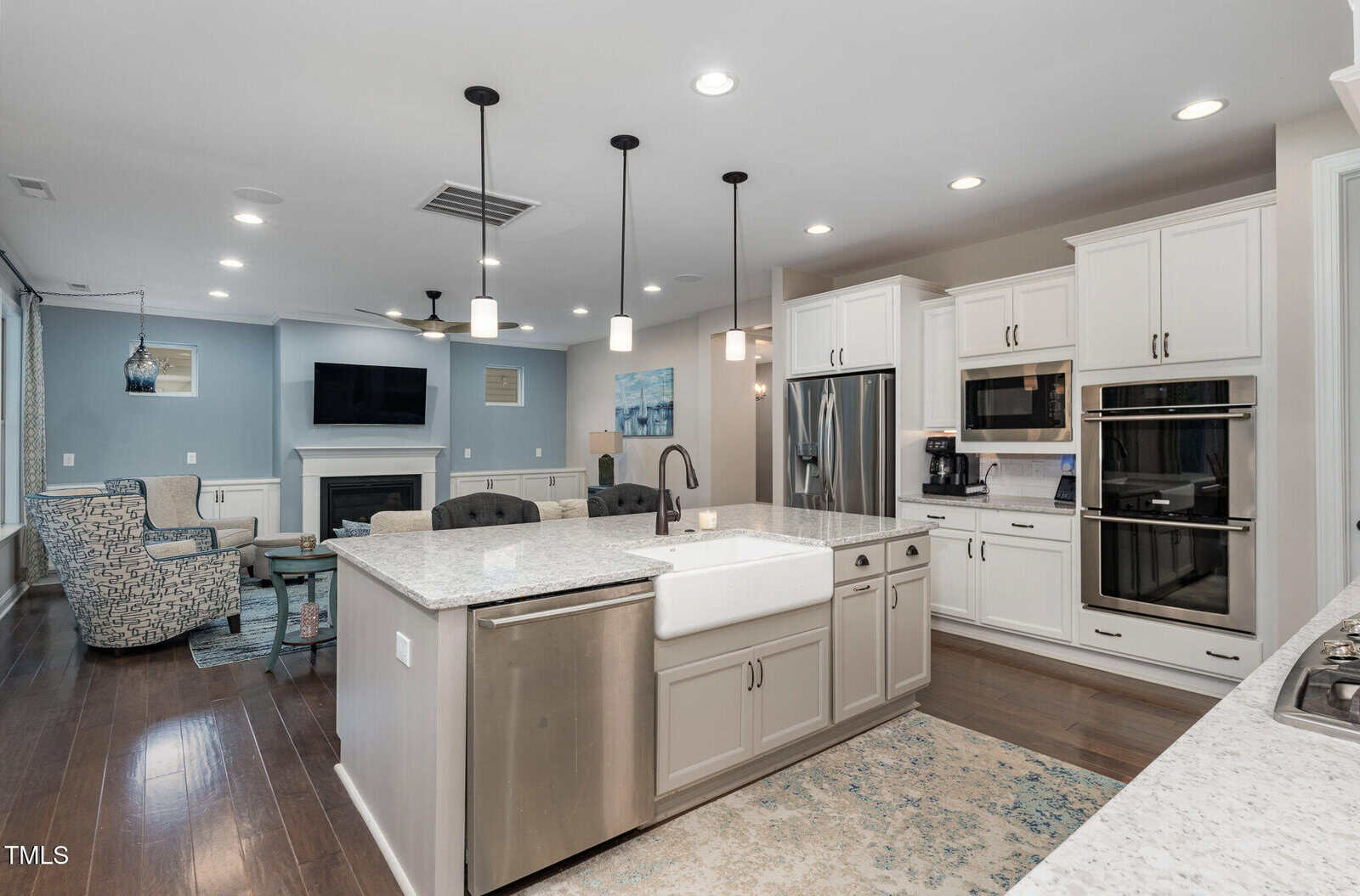 Modern kitchen with white cabinets, stainless steel appliances, a farmhouse sink, large island with seating, and open layout extending to a living area with a fireplace, TV, and armchairs. Bright, airy, and contemporary design.
