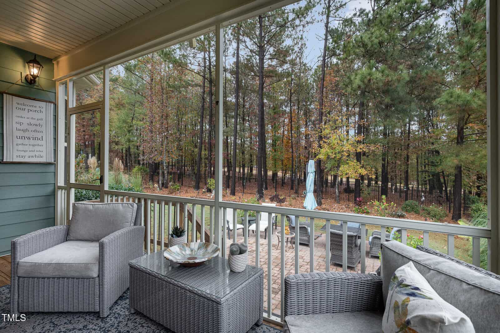 A screened-in porch with gray wicker furniture overlooks a patio with outdoor seating and a wooded backyard. The porch has a rug, cushions, and a decorative tray on the table. The sky is overcast.