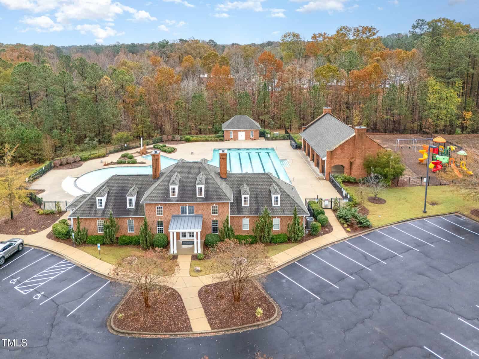 A brick clubhouse with a pitched roof sits in front of a swimming pool and a fenced playground area, surrounded by trees and bordered by a parking lot with several empty spaces.
