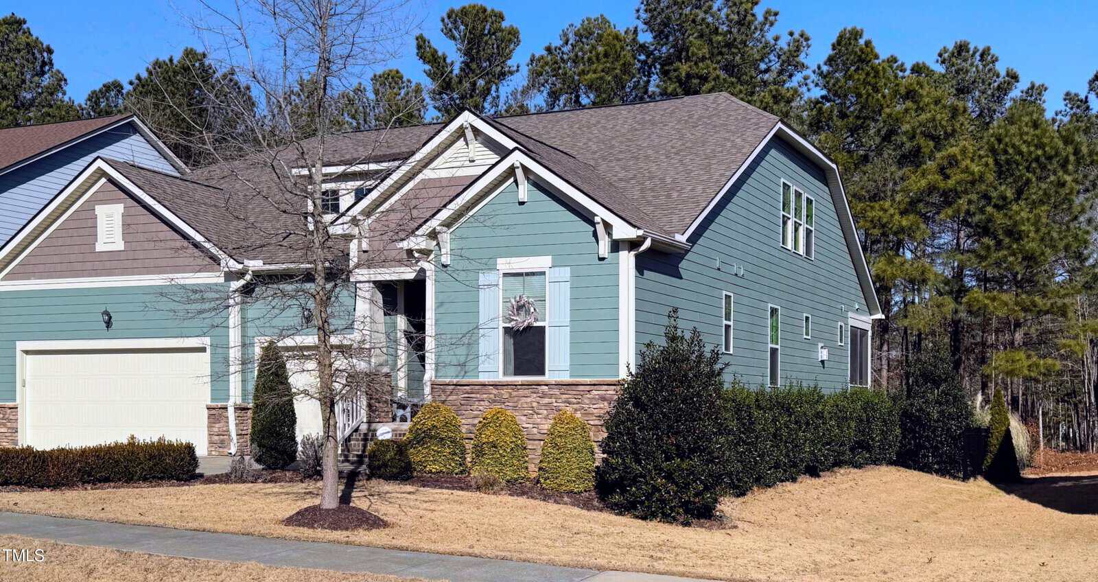 A two-story house with blue-green siding, stone accents, and white trim. There is a two-car garage, a small tree and shrubs in the front yard, and woods in the background under a clear blue sky.