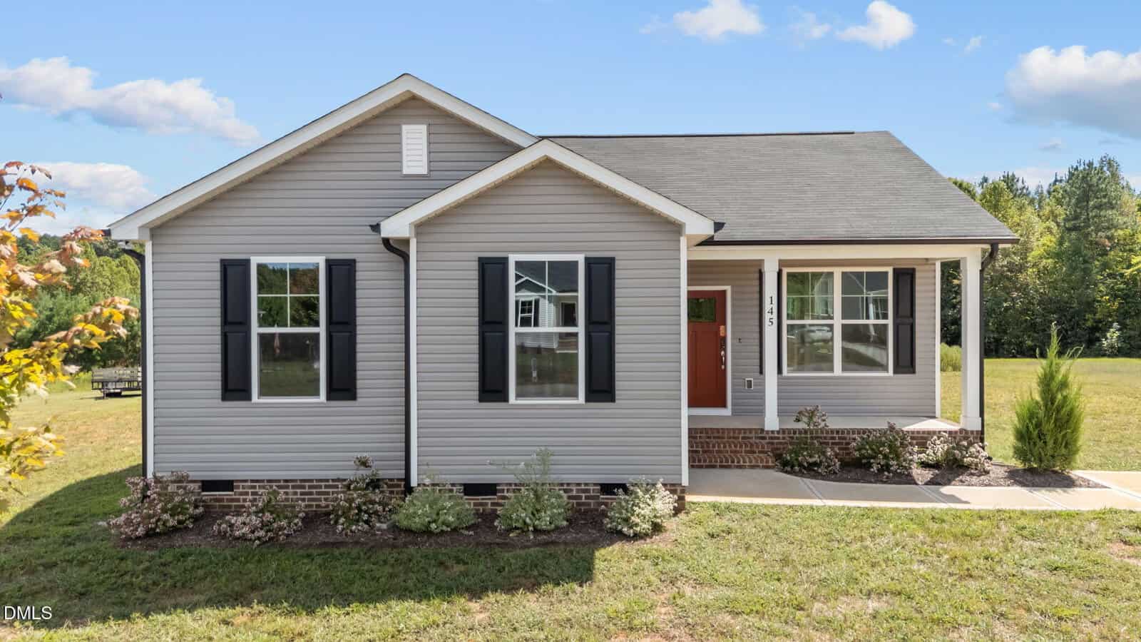 Charming single-story house at 145 Brookhaven Drive, Spring Hope, featuring gray siding, black shutters, a red front door, and a small covered porch. A few landscaped plants accent the front yard, while a classic brick foundation and sidewalk welcome you to the entrance. The home is surrounded by a lush grass yard for added curb appeal.