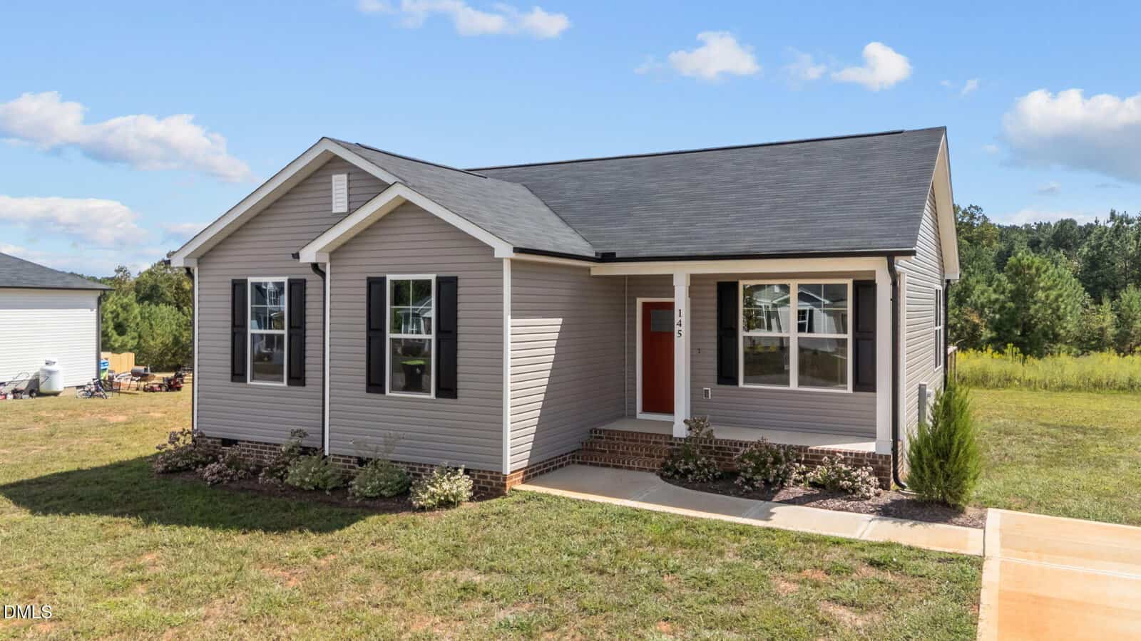 Single-story house with light beige siding, black shutters, a red front door, and a covered porch. The home has a small front yard with landscaping and a concrete walkway, set against a backdrop of trees and a partly cloudy sky.