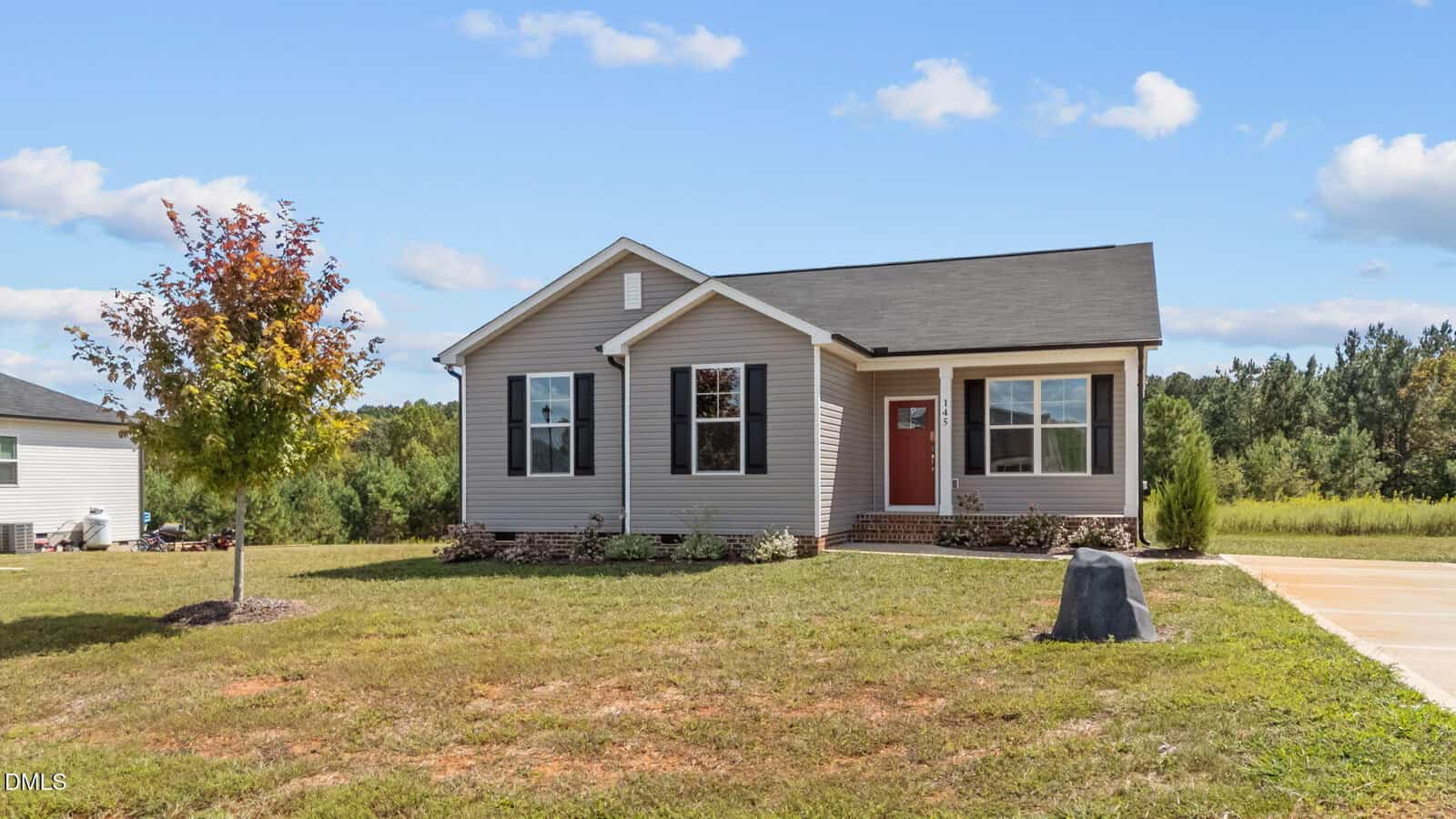 Single-story house with light siding, a black roof, and a red front door. The home has a small front porch, a young tree in the yard, a driveway, and is surrounded by grass and trees under a blue sky with clouds.