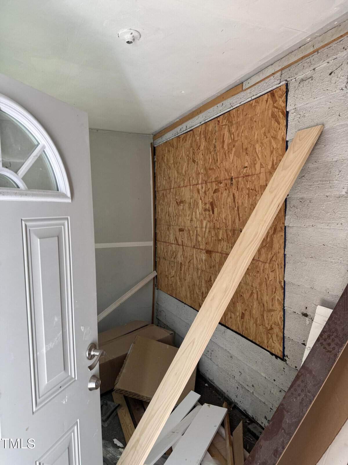 A cluttered room with an open door, showing building materials like plywood and wooden planks scattered on the floor. The walls are partially covered with wood panels. A beam leans against the wall.