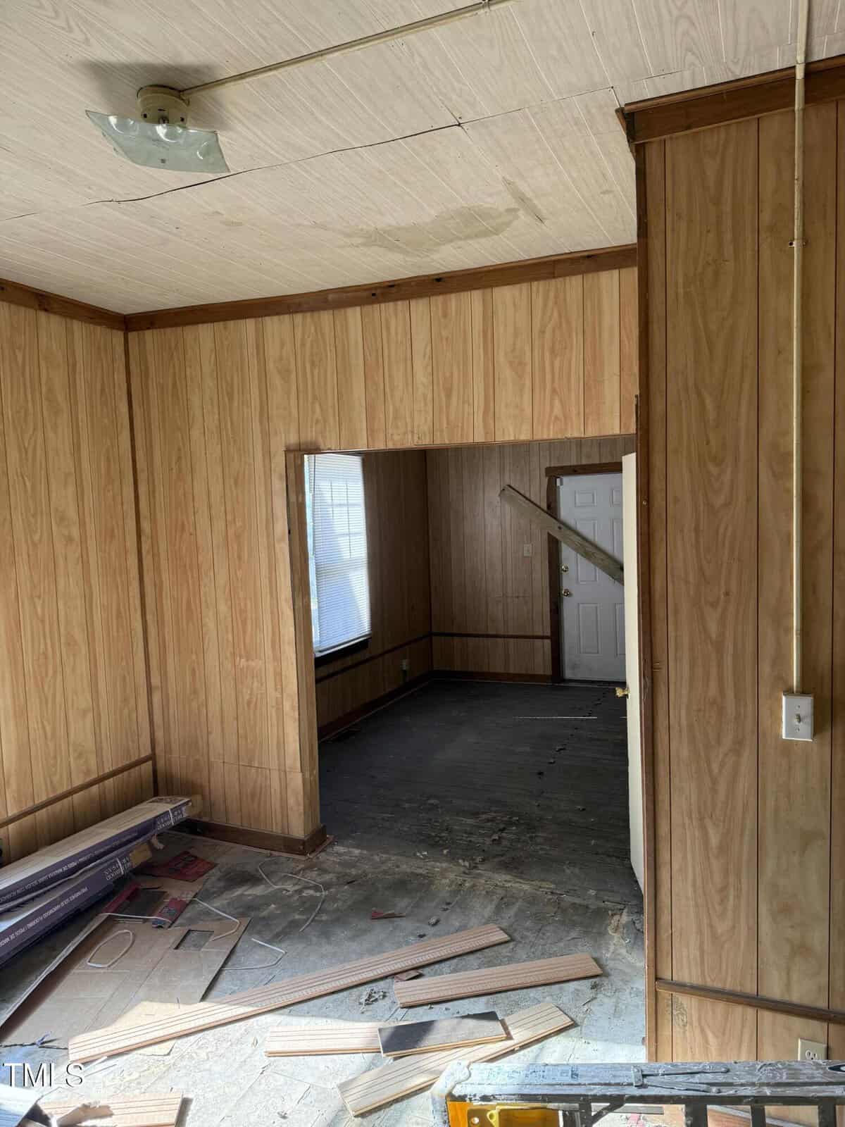 An empty room with wood-paneled walls and an unfinished floor. There's a light fixture on the ceiling and a window in the background. Pieces of wood and debris are scattered on the floor. An open doorway leads to another room.