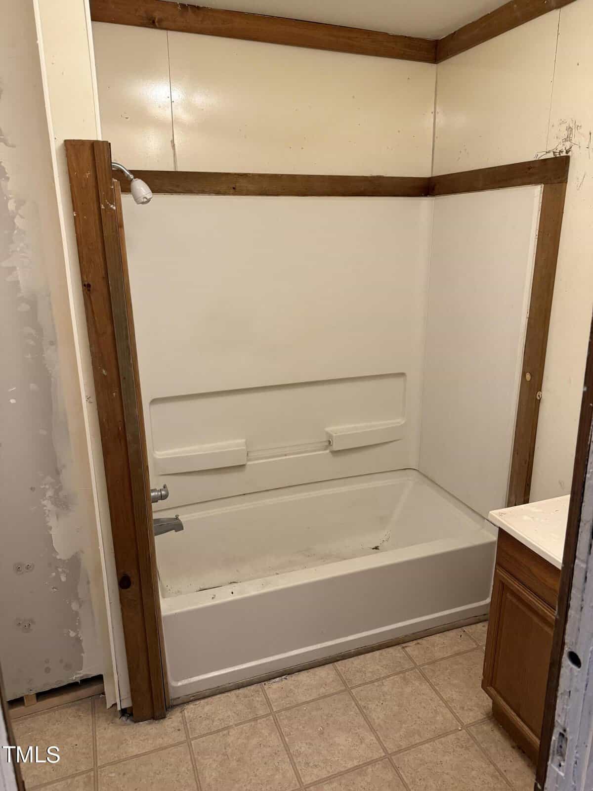 A small bathroom with a white bathtub and shower combination installed in a wooden-framed alcove. The walls are plain, and there is a partial view of a small vanity with a countertop on the right. The floor is tiled.