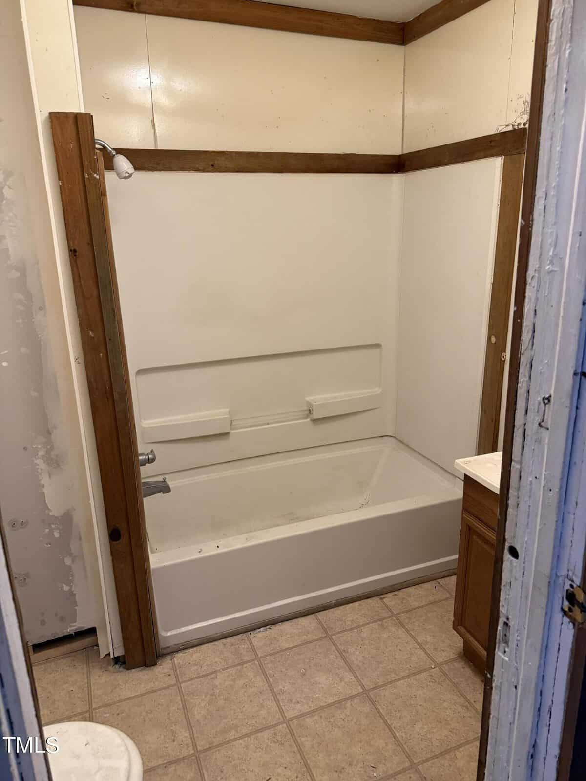 A small bathroom with a bathtub and showerhead, surrounded by beige walls and a wooden frame. The floor is tiled, and a wooden cabinet is partially visible on the right side. The room has a basic, unfinished appearance.