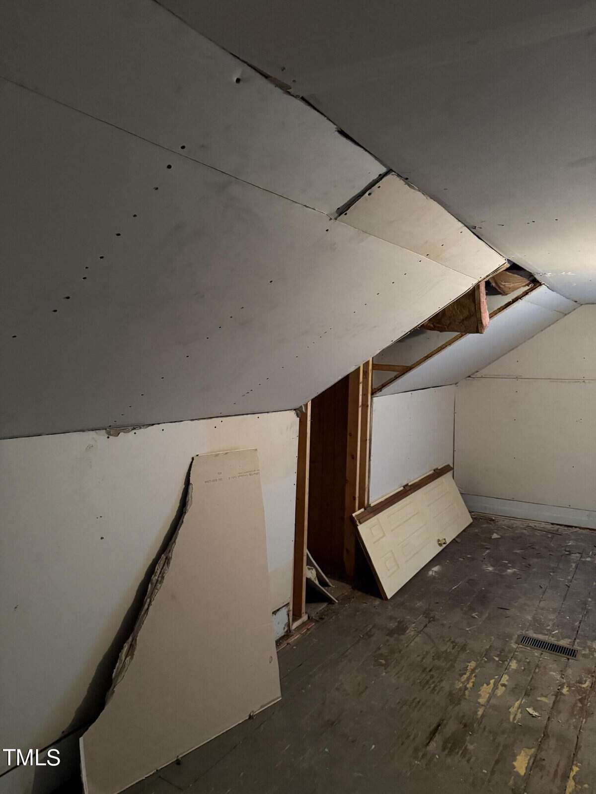 An attic space with partially installed drywall on the slanted ceiling. Some pieces of drywall are stacked against the wall, and a dismantled door is leaning on the right side. The floor appears unfinished or worn.