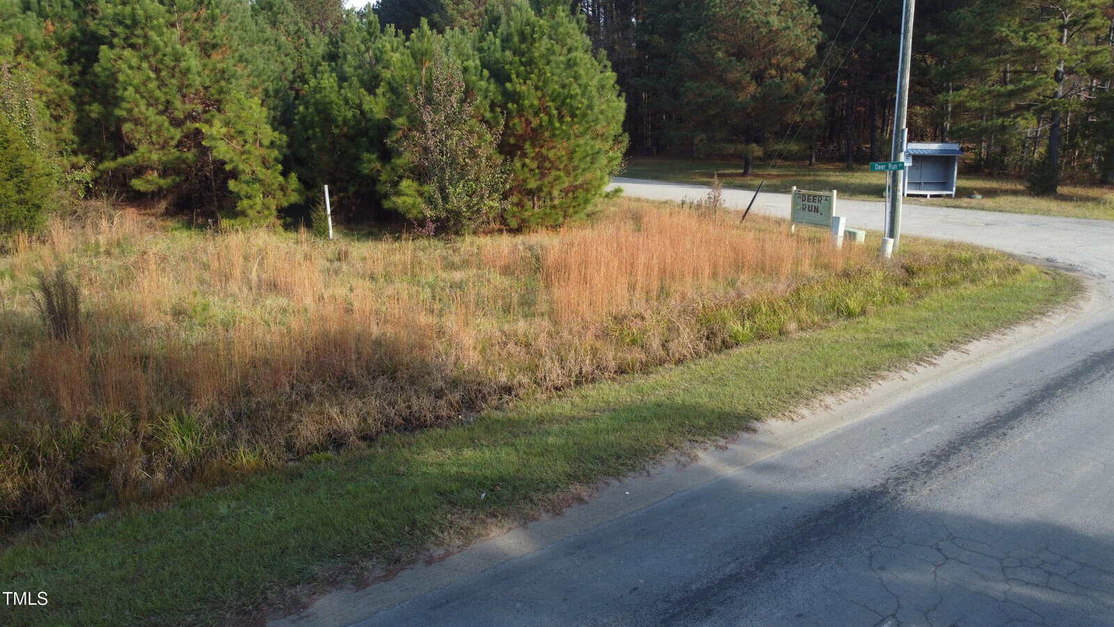 A rural intersection with a grassy roadside and a sign for a property called "Deer Creek Acres" near a cluster of trees. A small blue and white shelter is visible in the background, next to a paved road.