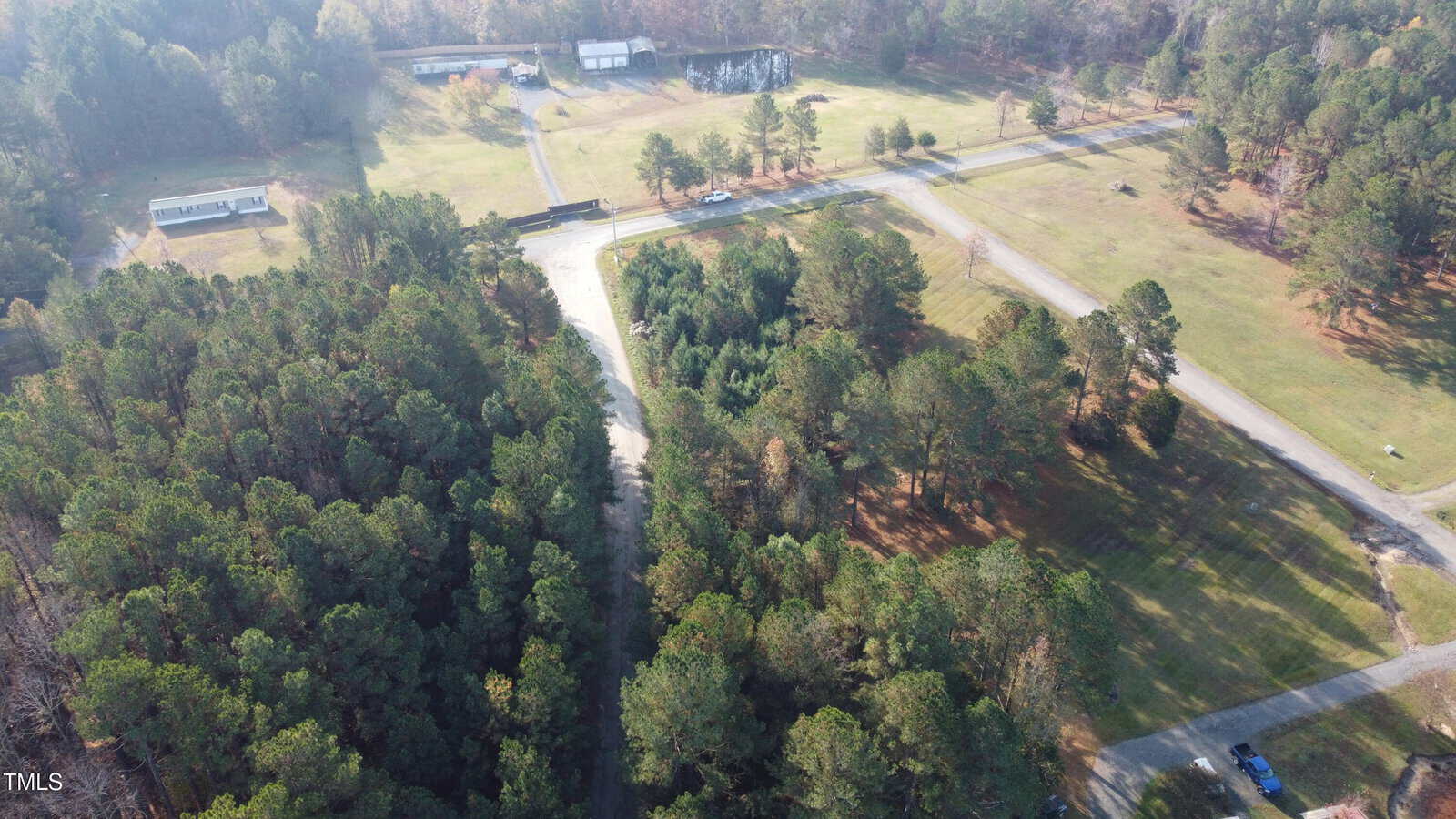 Aerial view of a rural landscape featuring a wooded area surrounded by open fields and roads. Buildings are visible in the background, with trees scattered throughout the scene. The lighting suggests a clear day.