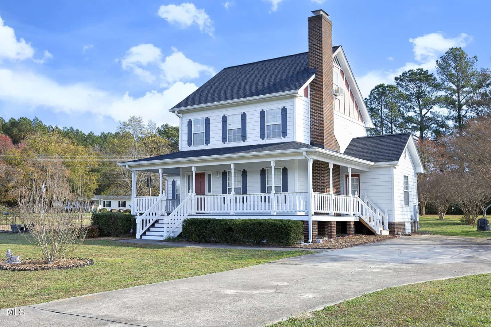 A two-story white house with a front porch, red shutters, brick chimney, and gabled roof sits beside a paved driveway. The yard has trees, shrubs, and a neatly edged lawn under a blue sky with scattered clouds.