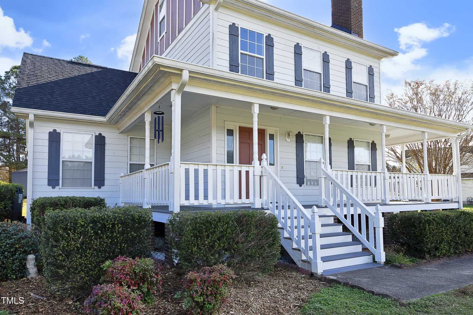 Two-story house with white siding, dark gray shutters, and a wraparound porch. The porch has white railings and steps, and there are neatly trimmed bushes and landscaping in front. The sky is partly cloudy.