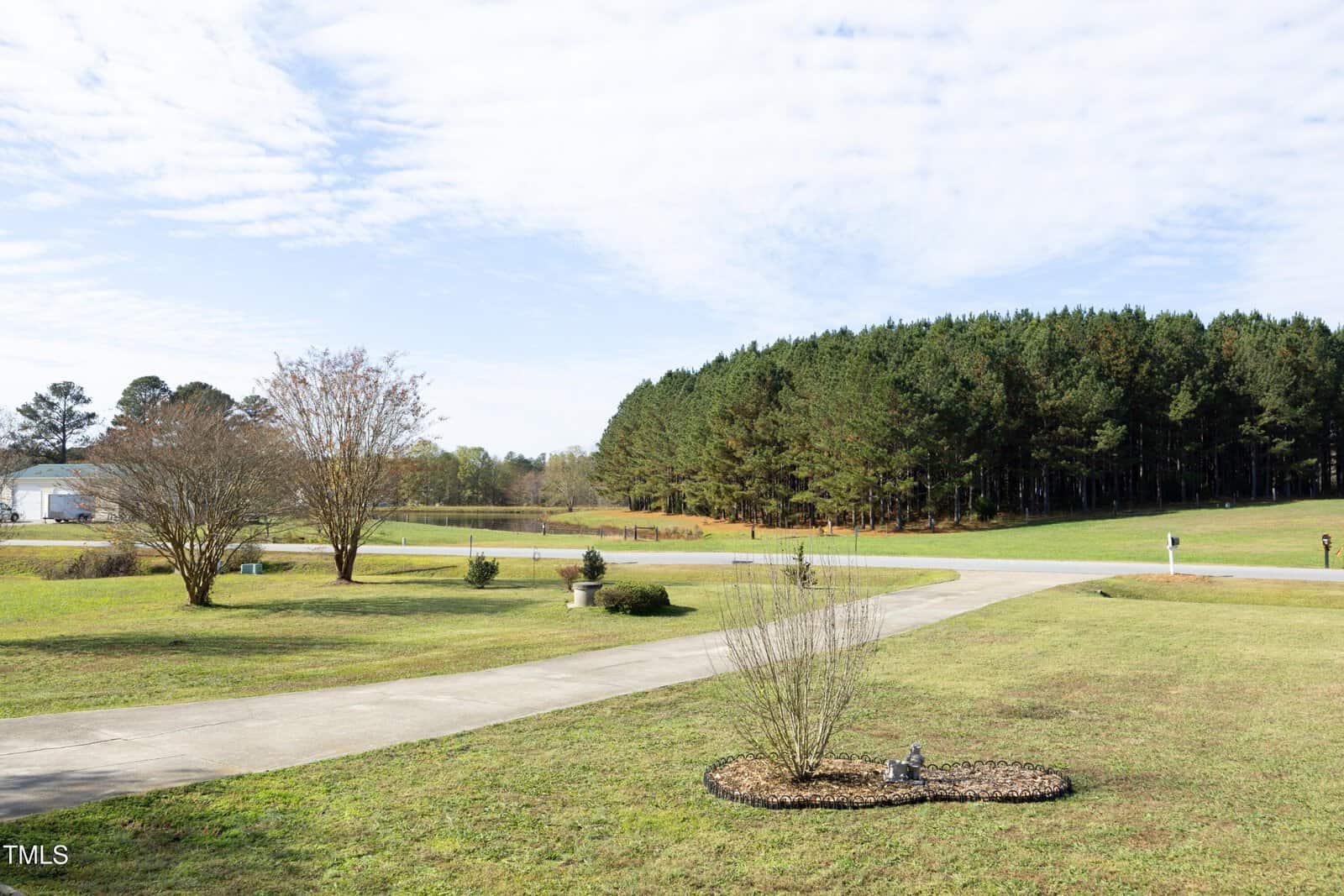 A curved driveway runs through a large, neatly trimmed lawn with a small landscaped area in the foreground. Tall trees line the background beneath a partly cloudy sky.