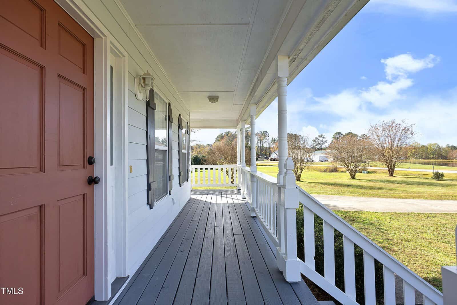 A covered front porch with white railings and posts, gray wooden flooring, and a red front door overlooks a driveway and grassy yard with trees in the background under a blue sky.