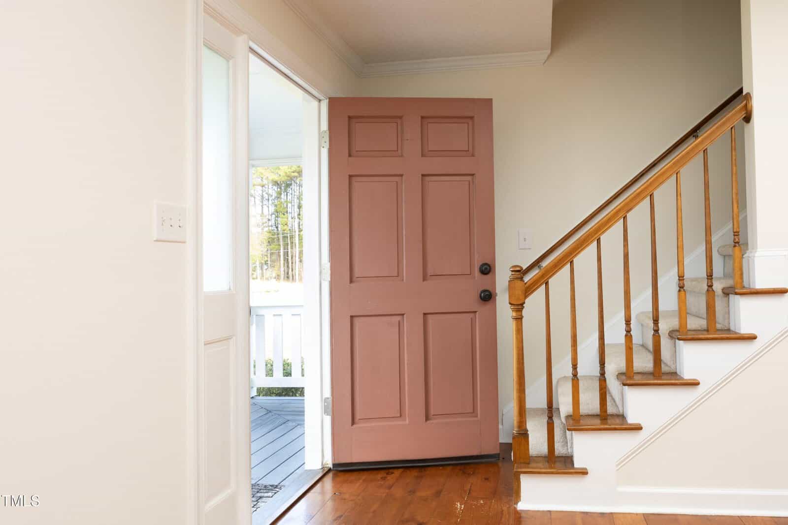 A wooden front door painted pink is open, revealing a white porch outside. To the right is a staircase with wooden handrails and carpeted steps. The interior walls are light-colored, and the floor is wood.