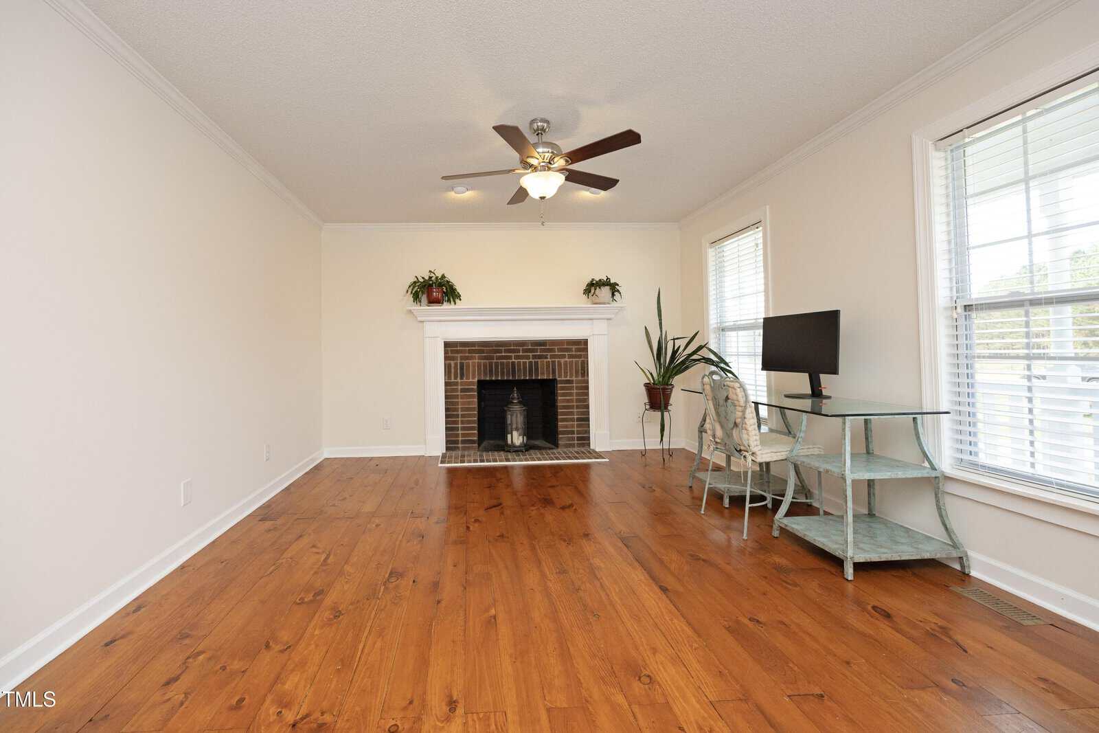 A bright living room with wood floors, a brick fireplace with a white mantel, two potted plants, a ceiling fan, and a glass-topped desk with a computer monitor beside windows with white blinds.