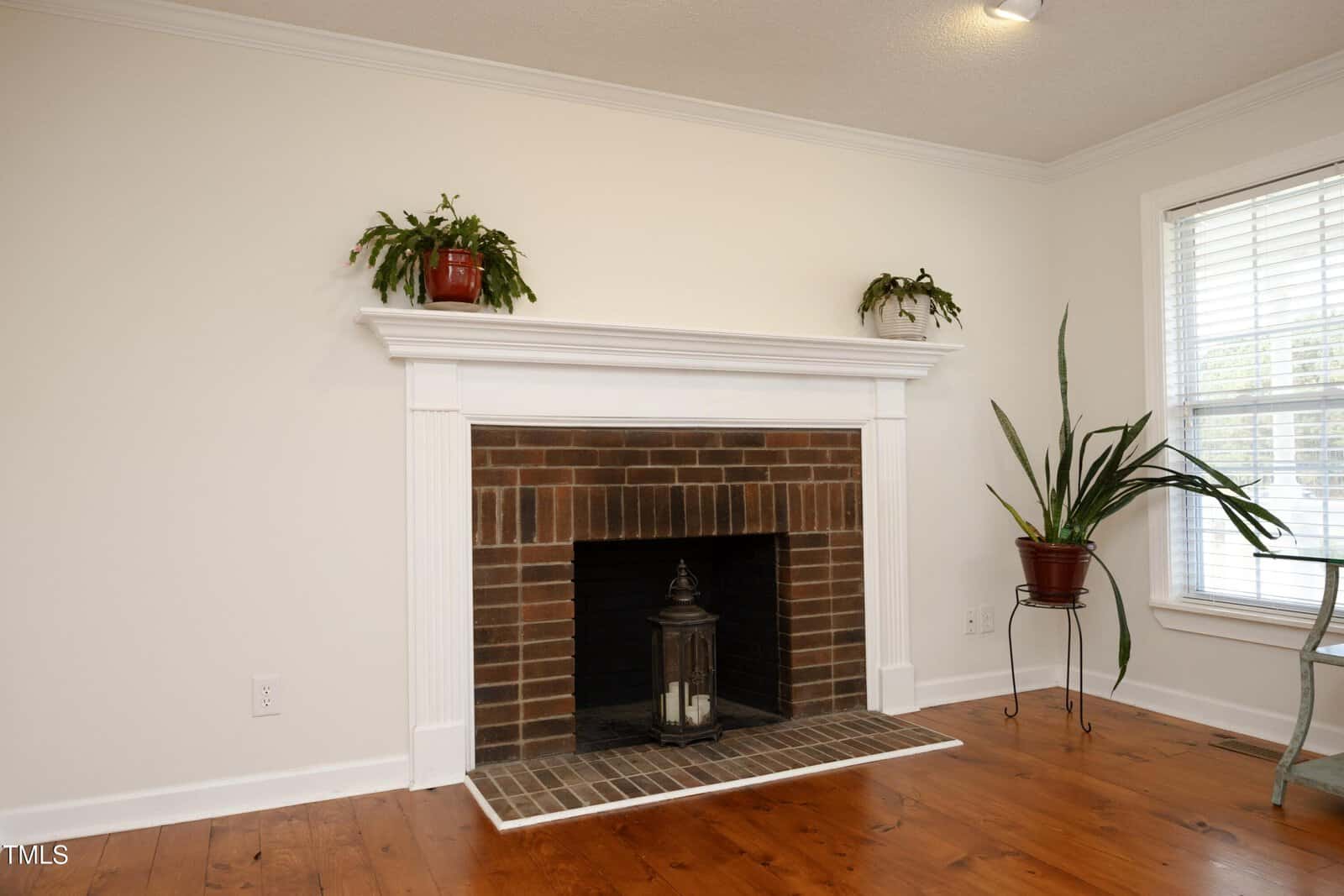 A white mantel fireplace with a brick interior, a large lantern with candles inside the hearth, two potted plants on the mantel, and a tall potted plant on a stand to the right. The room has white walls and wood flooring.