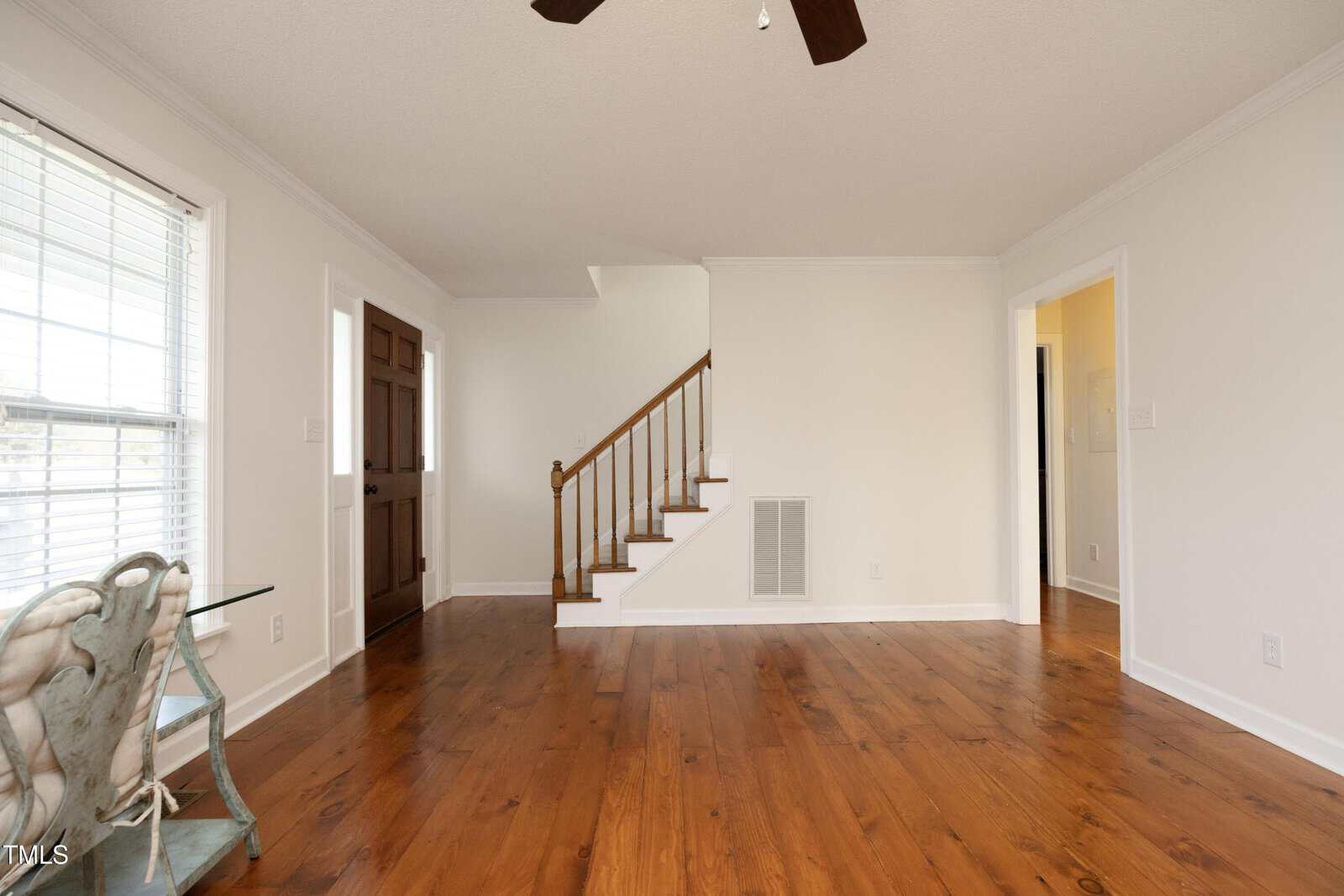 A bright living room with wooden floors, white walls, a staircase with a wooden handrail, a brown door, large windows, and an ornate chair in the corner.
