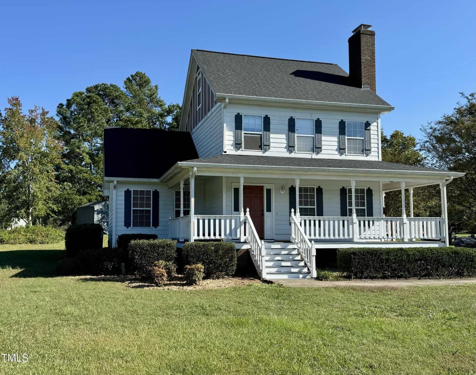 Two-story white house with a large front porch, black shutters, a red front door, and a brick chimney, surrounded by hedges and grass under a clear blue sky.