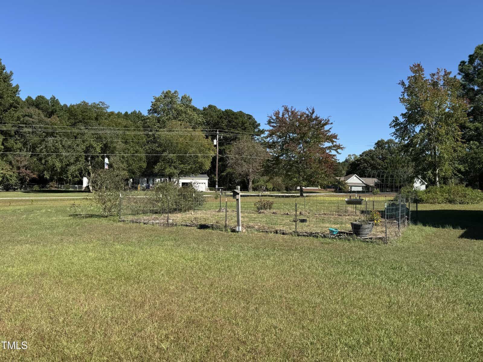 A fenced garden with plants and gardening tools sits in a grassy yard, bordered by trees and a road with power lines. Houses and clear blue sky are visible in the background.