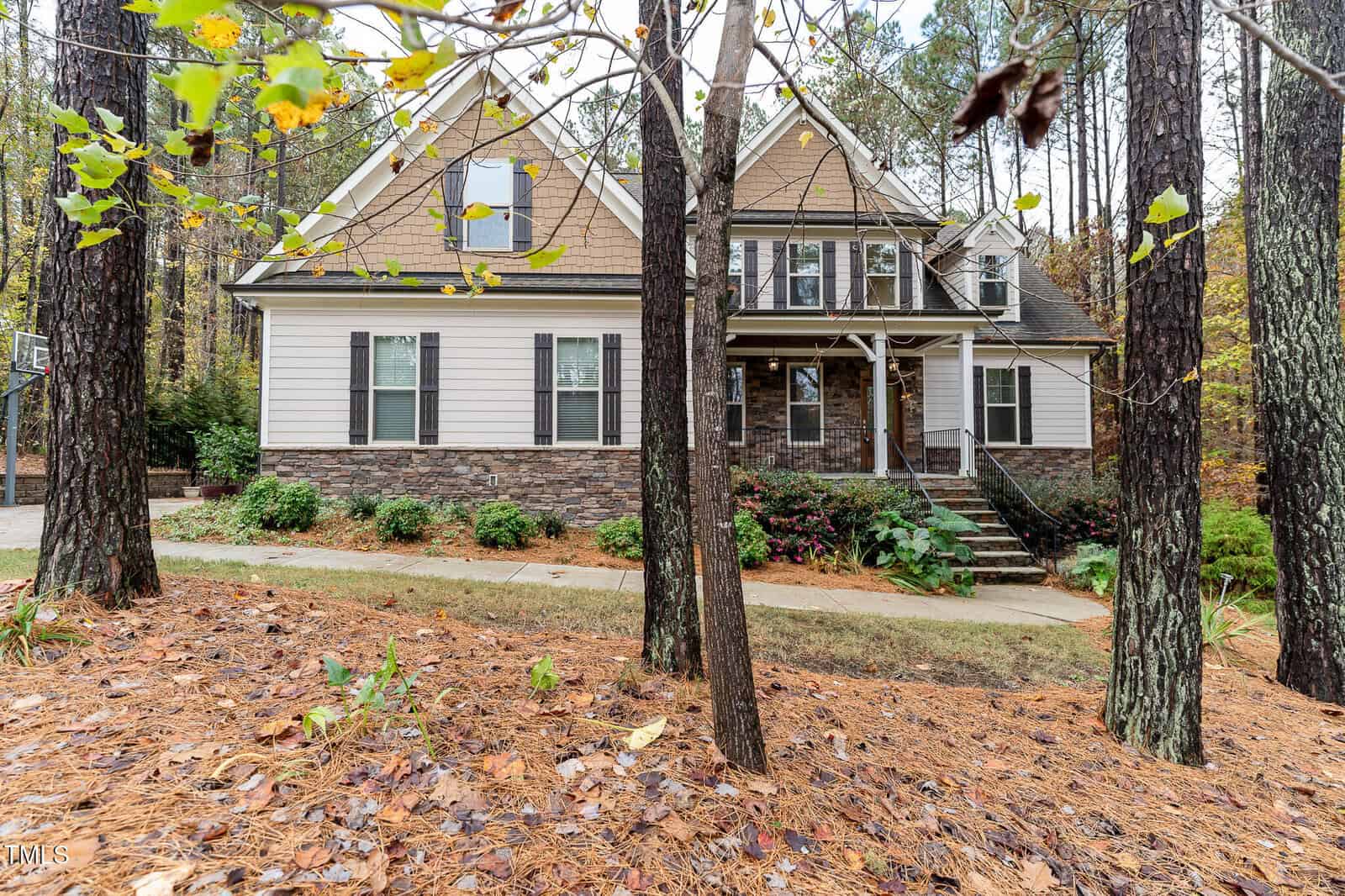 Nestled in Wake Forest, the two-story house on 3584 Claude Court showcases a blend of stone and beige siding, surrounded by trees. The front boasts a small porch with steps, a gabled roof, and multiple windows. Fallen leaves blanket the ground, capturing the essence of autumn.