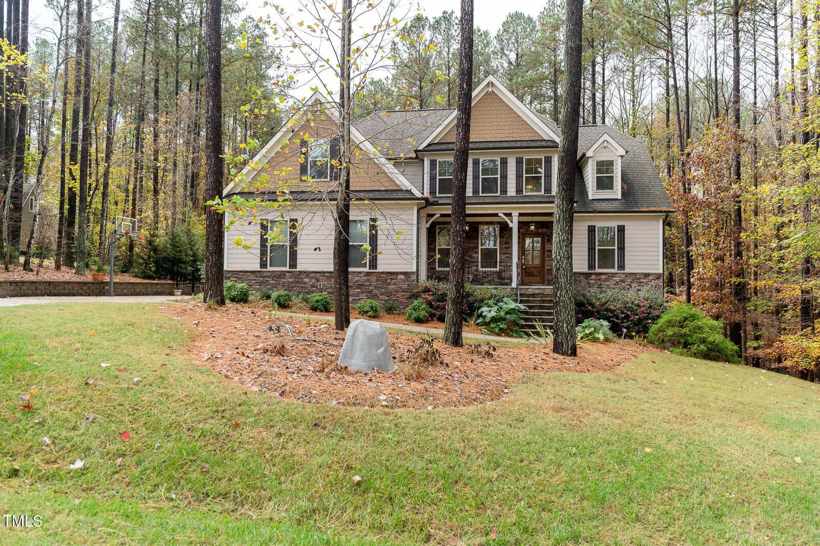 A two-story house with a combination of stone and siding exterior is set among tall trees. The front yard is landscaped with a circular bed of mulch and plants. The driveway curves around the left side of the property.