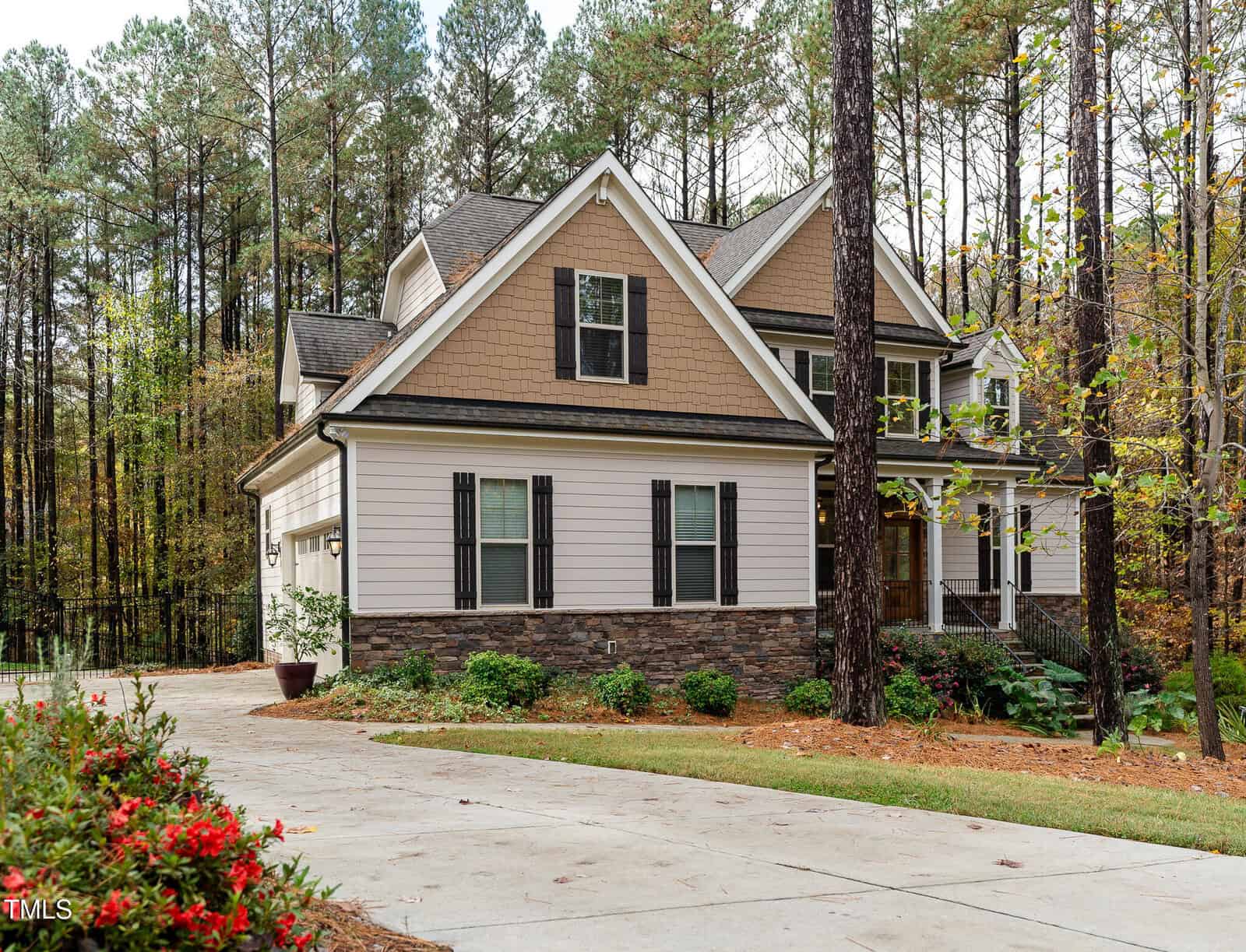 A two-story house with beige siding, white trim, and stone accents is surrounded by tall pine trees. The driveway curves to the left, and there are red flowers in the foreground. A potted plant sits near the left of the house's entrance.