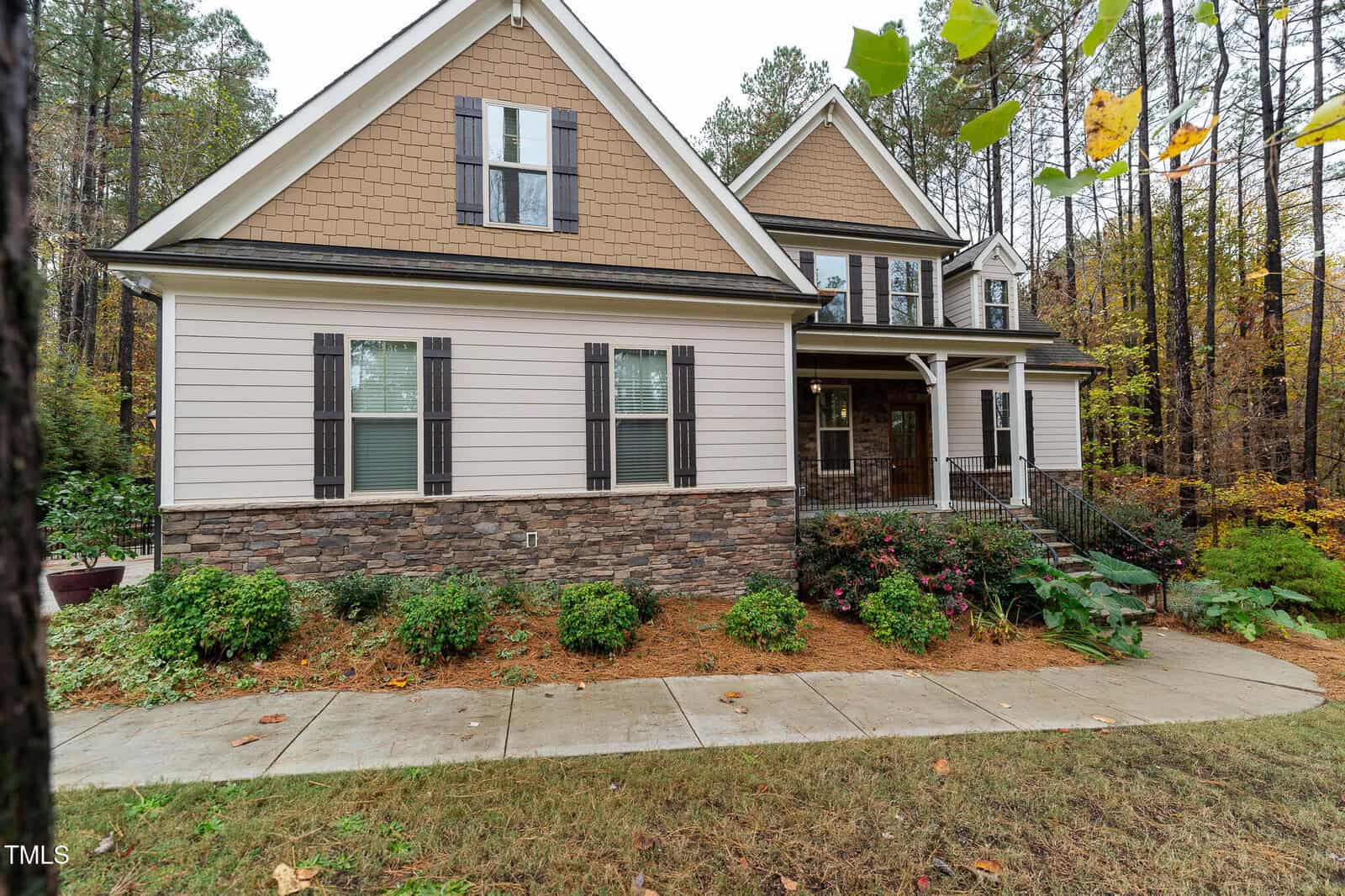 A two-story house with light-colored siding, stone accents, and dark shutters, set against a backdrop of tall trees. The front features a small porch with railings and a landscaped yard with shrubs. A concrete walkway leads to the entrance.